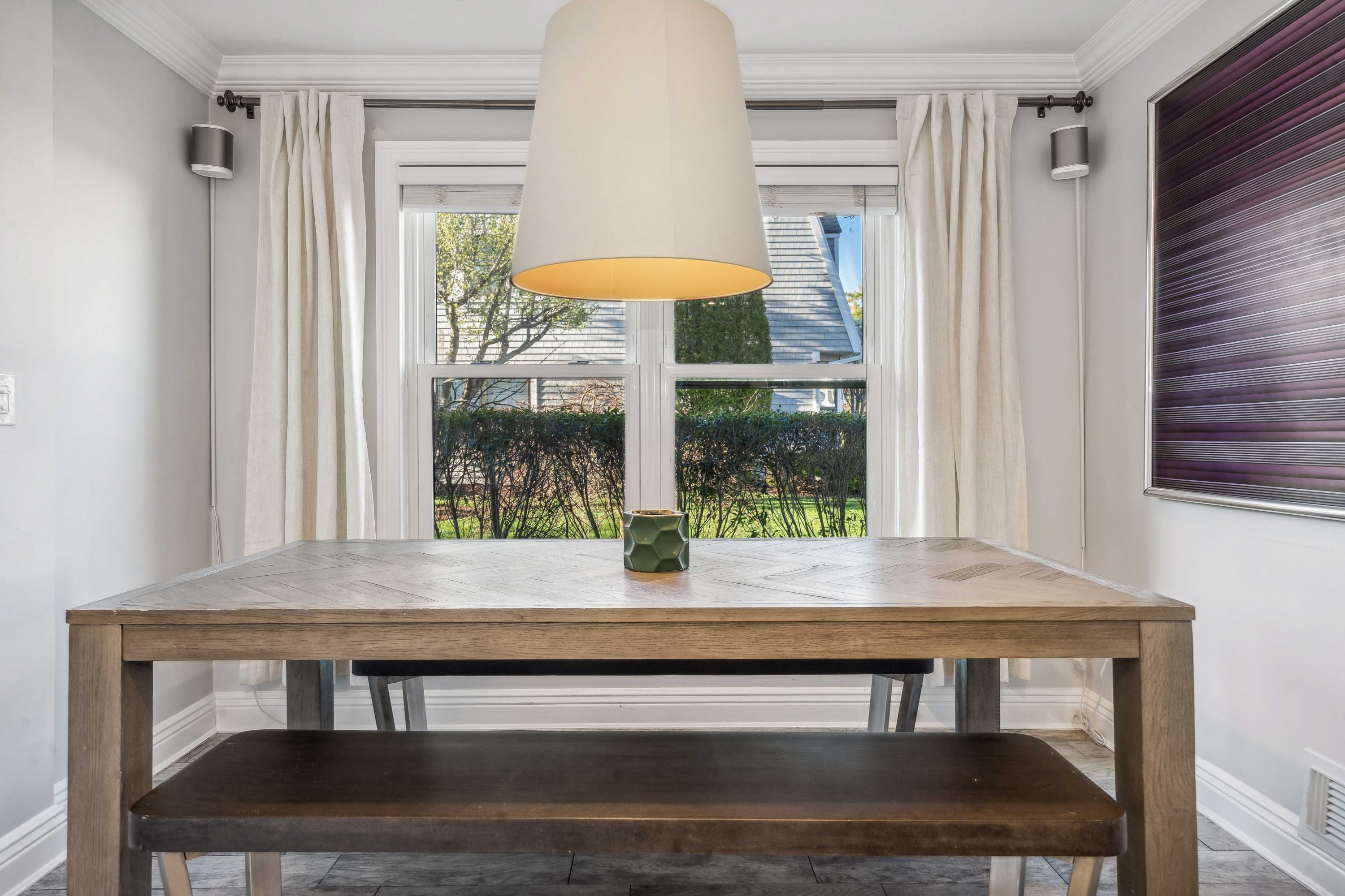 Dining area with a wood table and bench set beneath a linen pendant light, near a bay window.