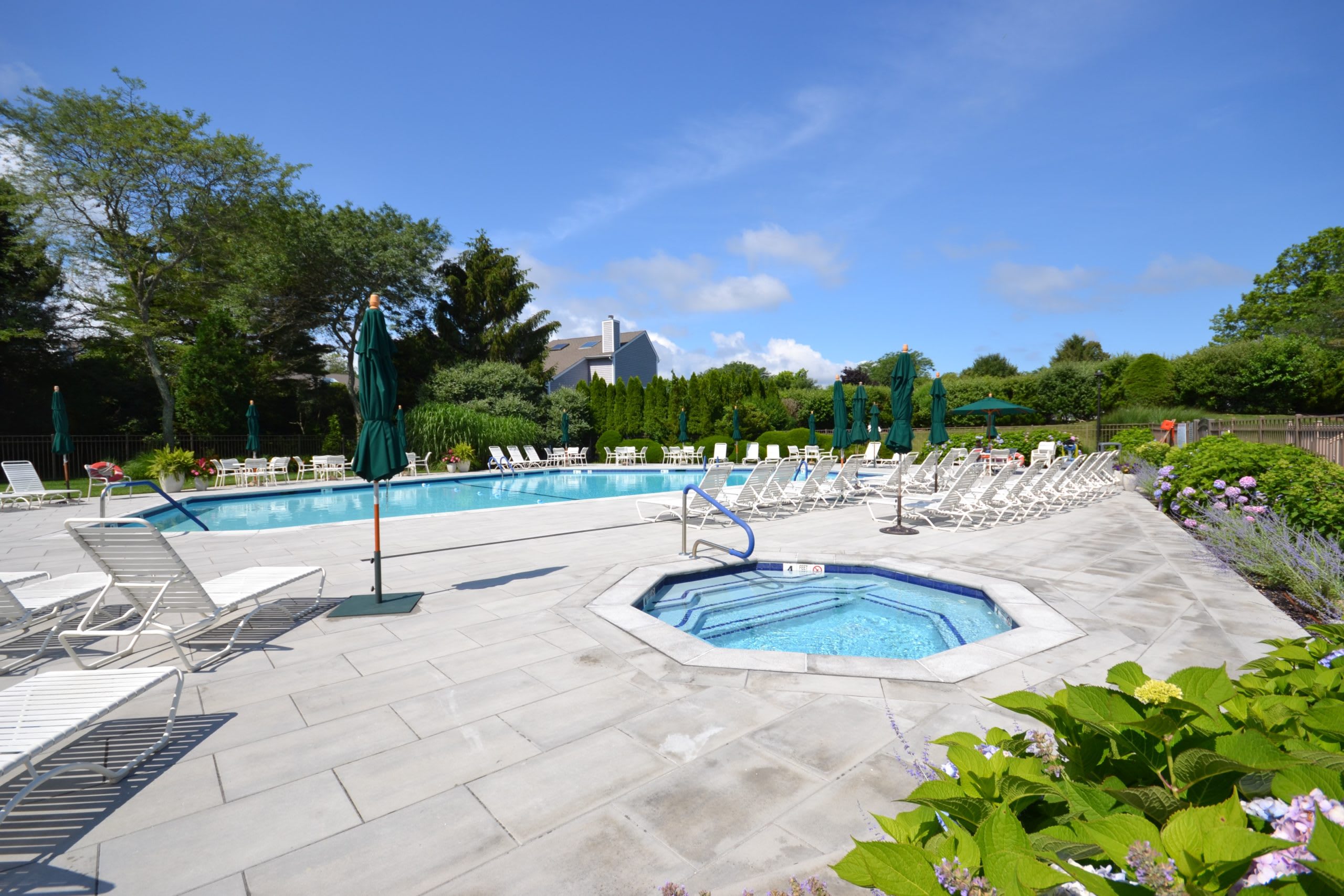 Community pool and spa with lounge chairs on a stone patio surrounded by greenery.