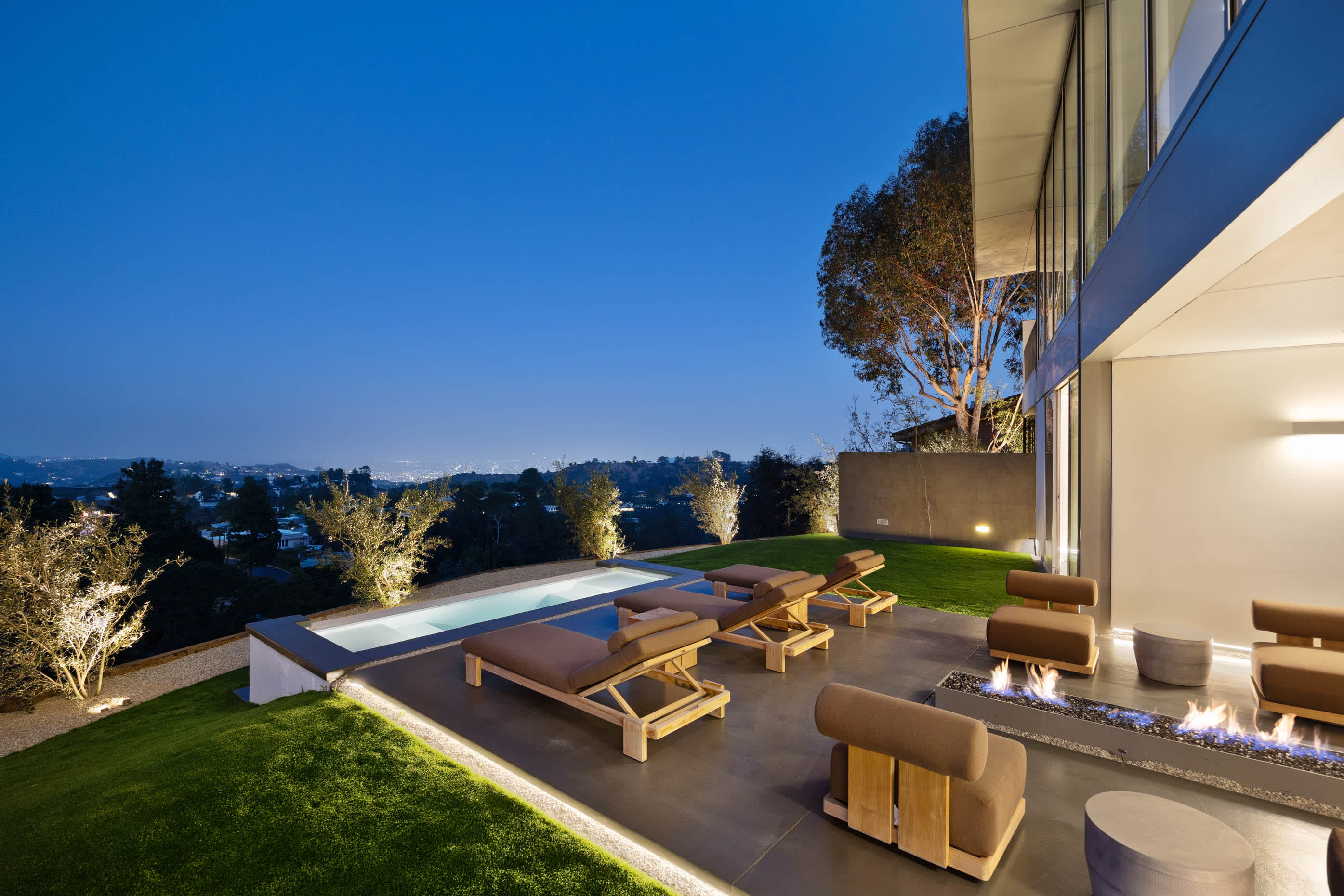 Outdoor space with loungers, a night-lit pool, a fire pit, and luxurious seating, all overlooking the city.