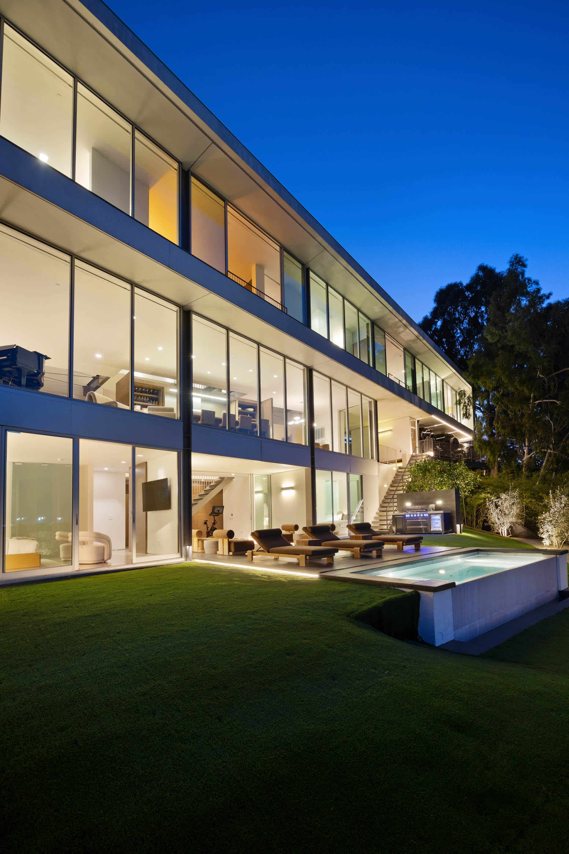 Three-story architectural marvel with expansive glass walls, a sleek pool, stylish loungers, and a meticulously maintained yard.