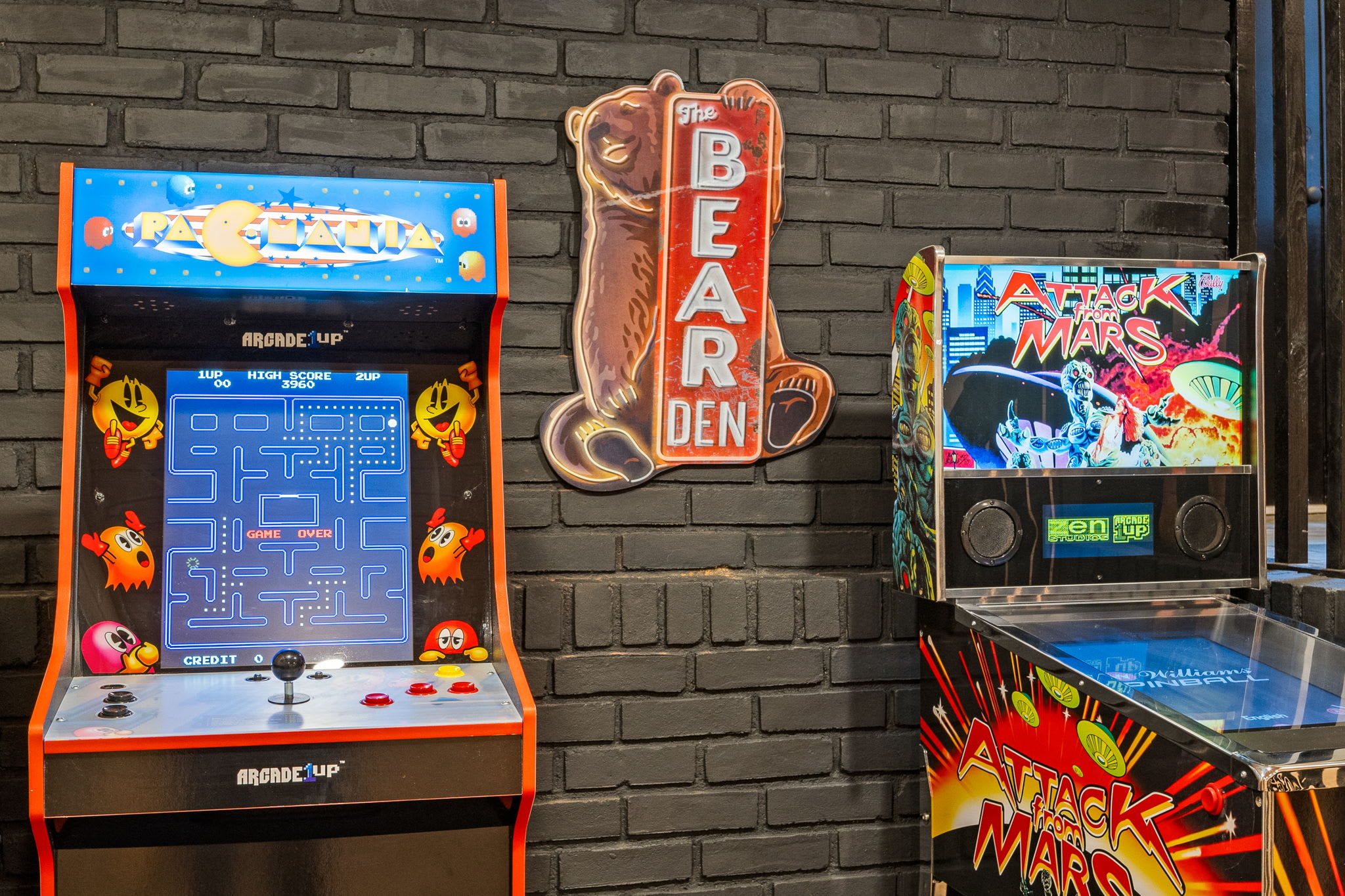 Classic Gaming Den – The Bear Den – A fun and lively arcade area with vintage Pac-Man and pinball machines, accented by a bold brick wall and neon sign.