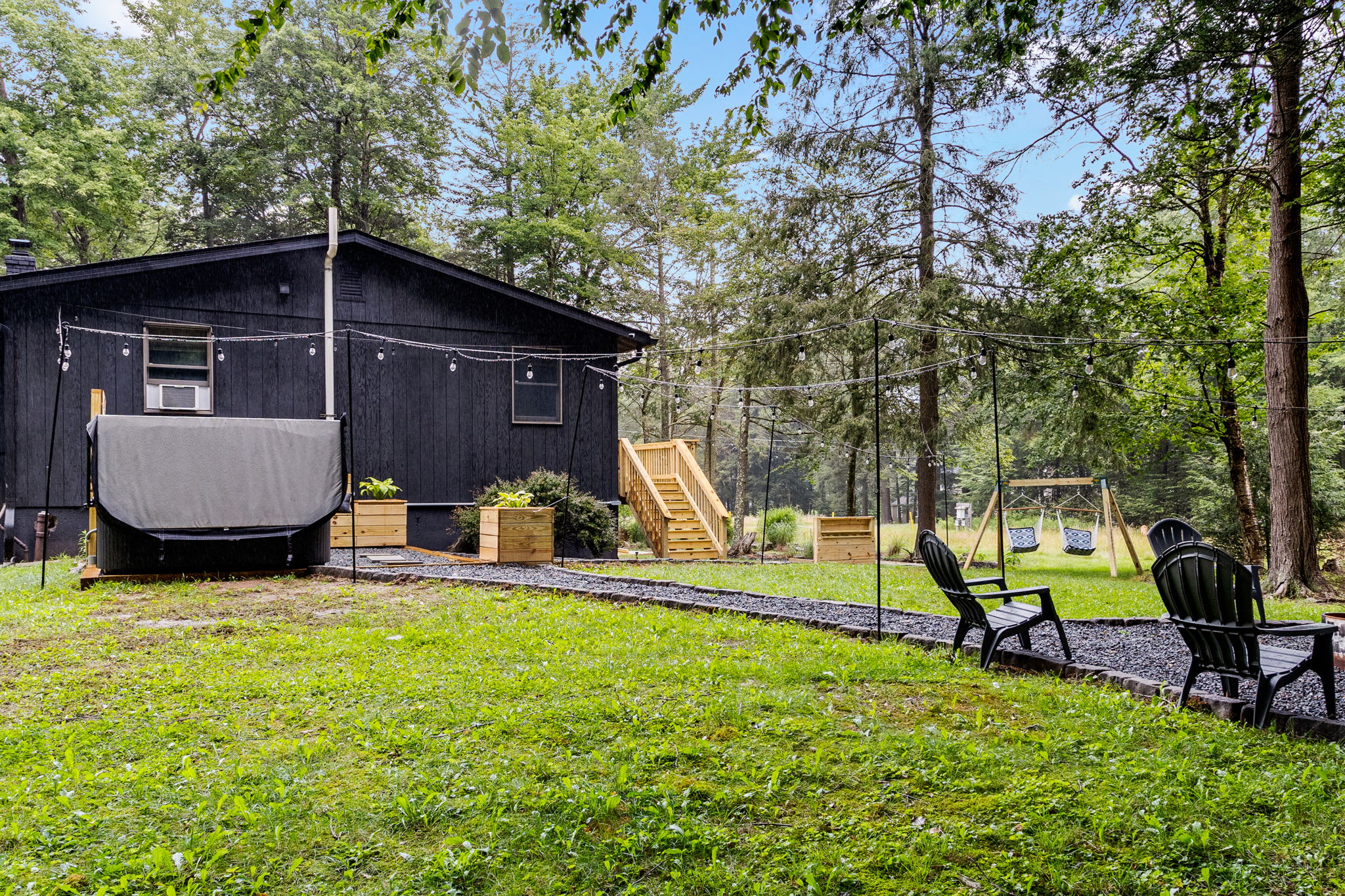 A modern black cabin nestled in the woods, complete with a fire pit, hot tub, and lush greenery for the ultimate escape.