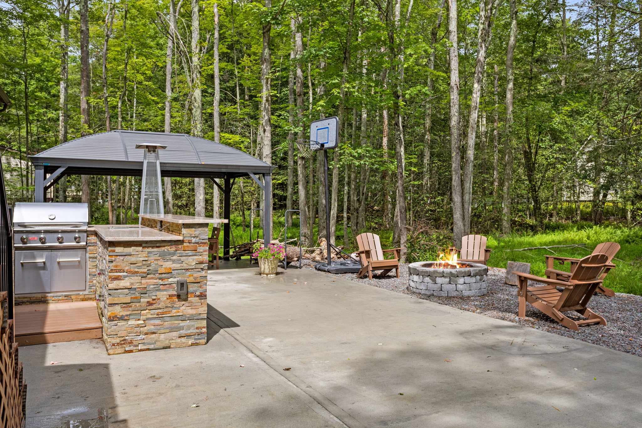 Escape to this serene outdoor haven featuring a firepit, a full outdoor kitchen, and a basketball hoop, all surrounded by lush forest for the perfect nature getaway.