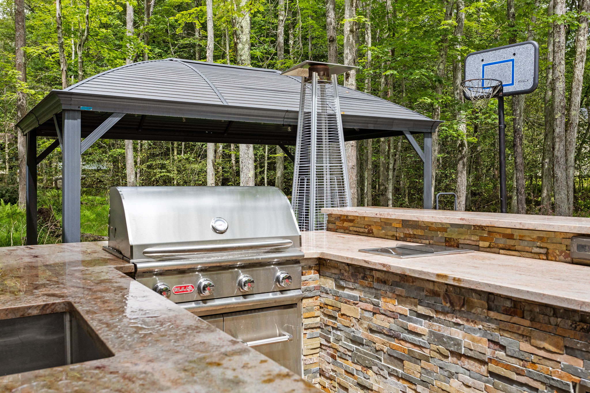 Enjoy the best of outdoor living with this stunning kitchen setup, featuring a stylish stone design, a covered dining space, and a cozy firepit nearby.