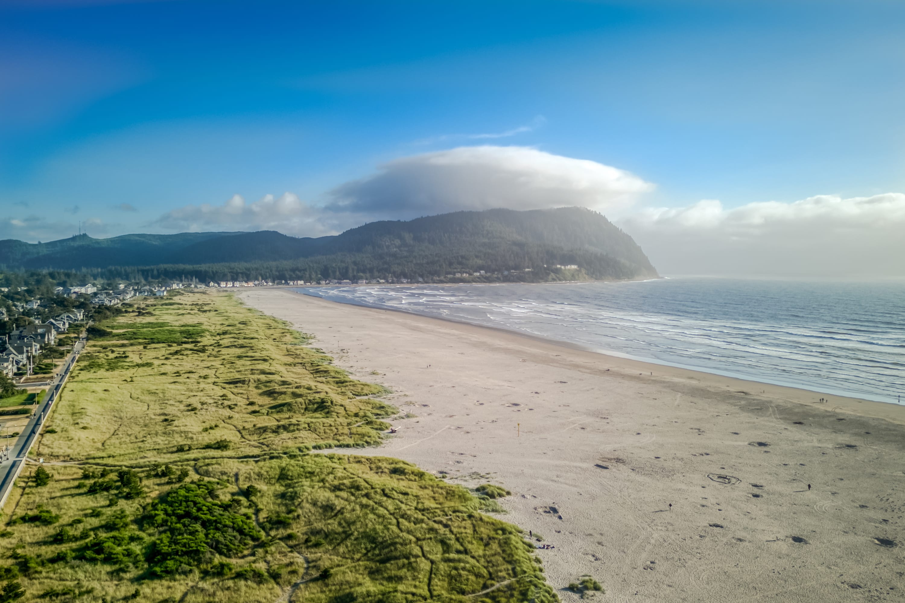 An expansive aerial view captures a wide sandy beach stretching along the coastline, bordered by green grassy dunes and a town with buildings. A large forested headland juts into the ocean under a partly cloudy blue sky.