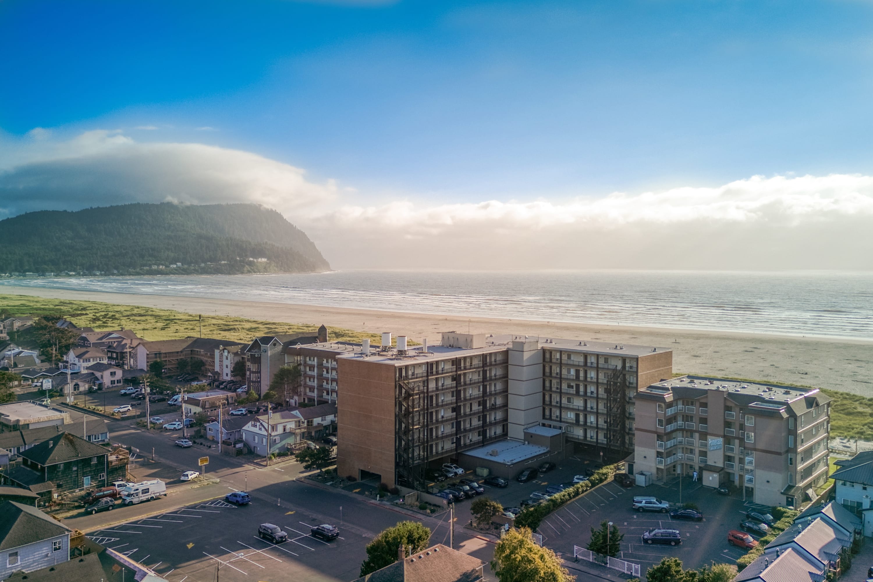 An aerial view captures a large oceanfront condominium complex with a parking lot, situated directly adjacent to a wide sandy beach. The ocean stretches to the horizon under a partly cloudy blue sky, with a large, cloud-covered mountain in the distance.