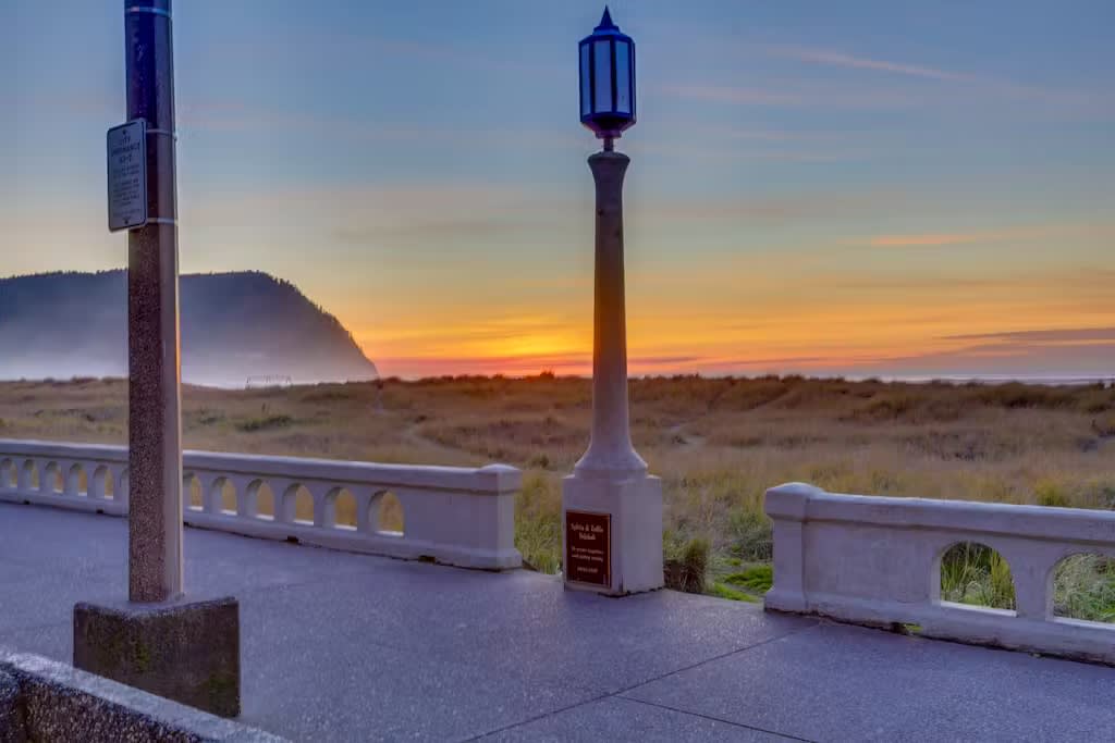 A paved promenade with elegant lampposts overlooks a serene beach scene at sunset, featuring grassy dunes and the ocean. The sky transitions from deep blue to warm orange and yellow, with a distant mountain shrouded in mist.