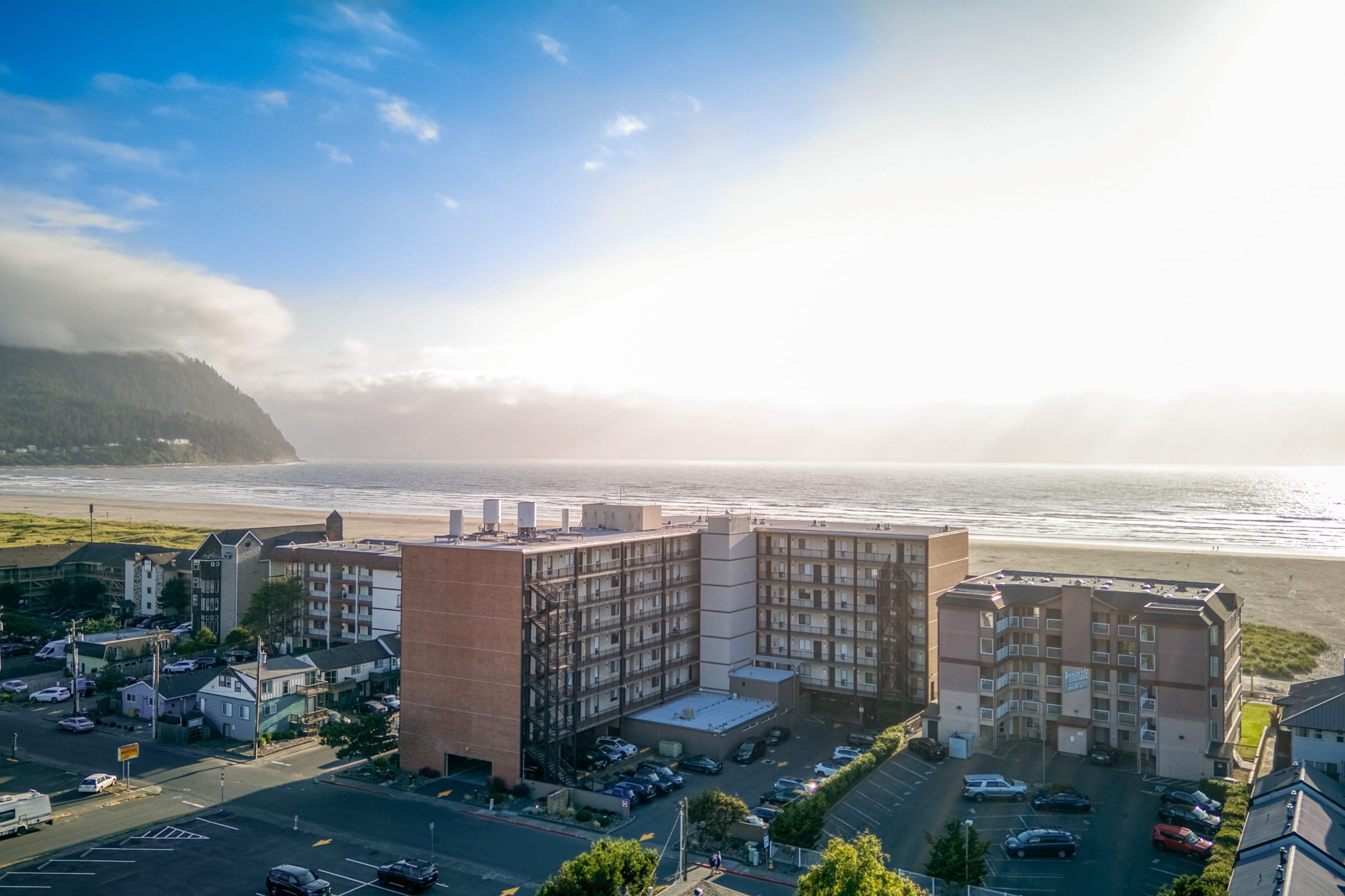 An aerial perspective showcases a multi-story brick and concrete condominium building with numerous balconies, situated directly across from a wide sandy beach and the ocean. Surrounding residential buildings and a parking lot are also visible under a bright, sunny sky.