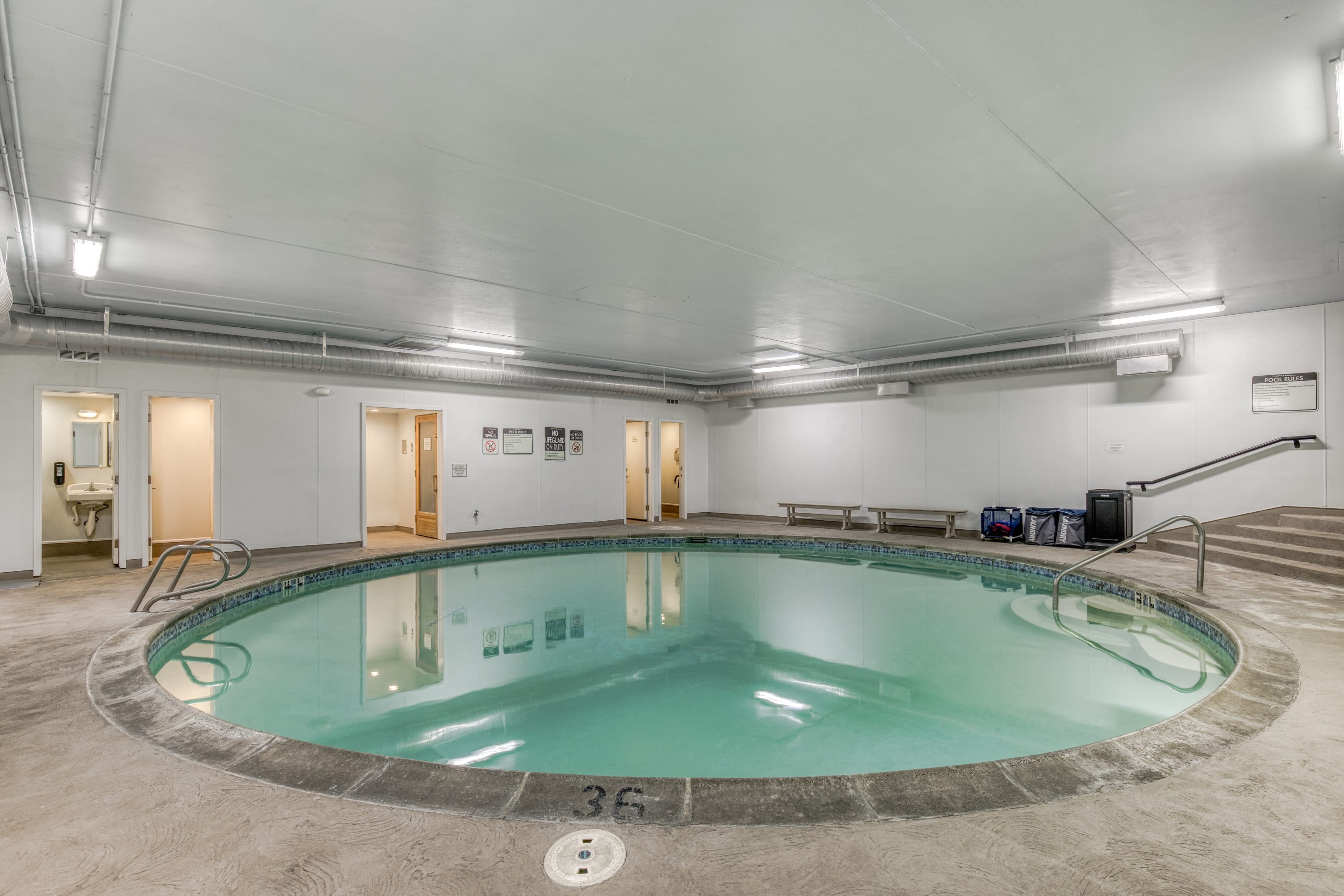 A clean, circular indoor pool with steps and handrails, set in a brightly lit room with concrete flooring and access to changing rooms.