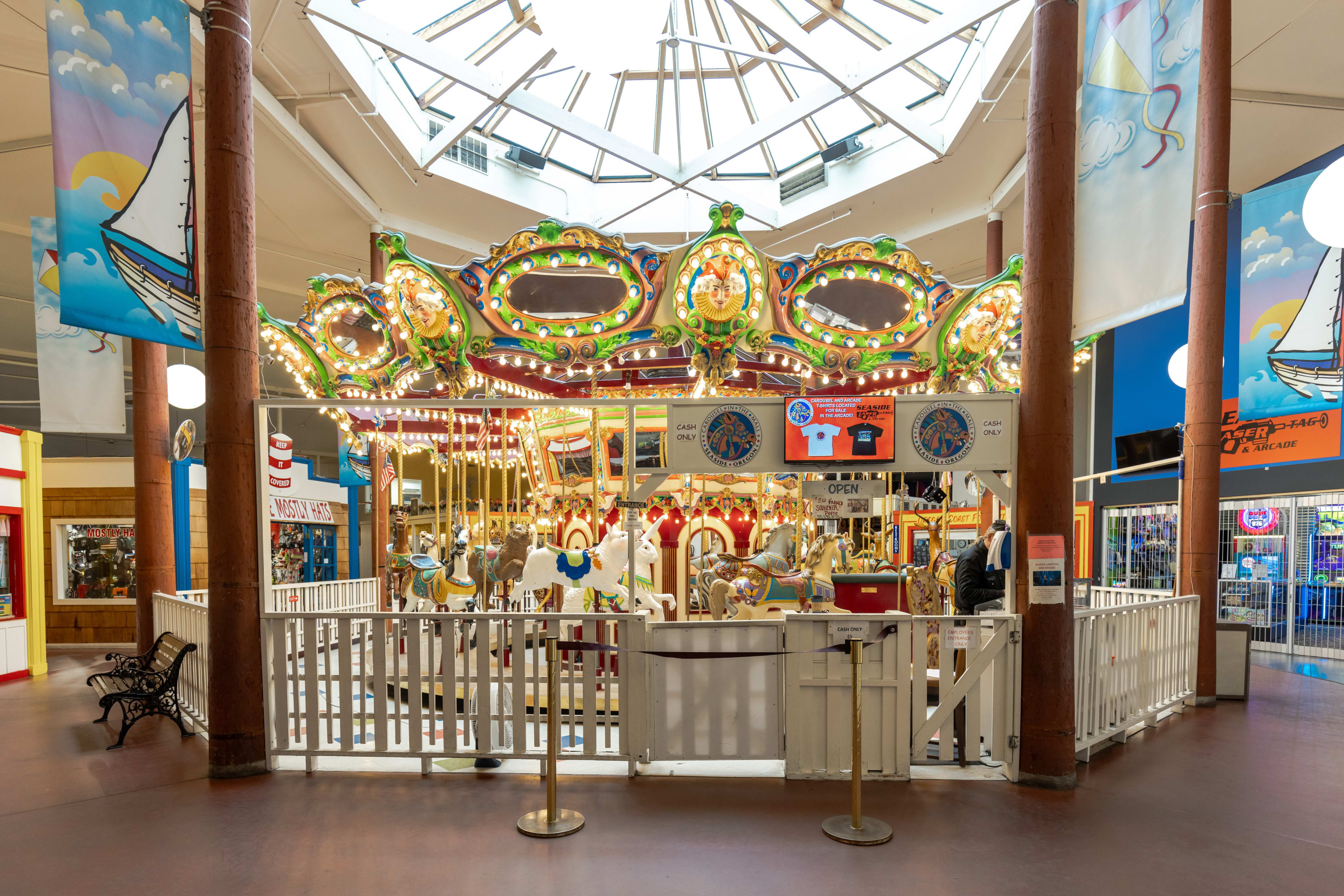 A brightly lit indoor carousel features numerous ornate horses and other animals, surrounded by a white fence and banners depicting sailboats. The ceiling has skylights, allowing natural light to illuminate the ride.