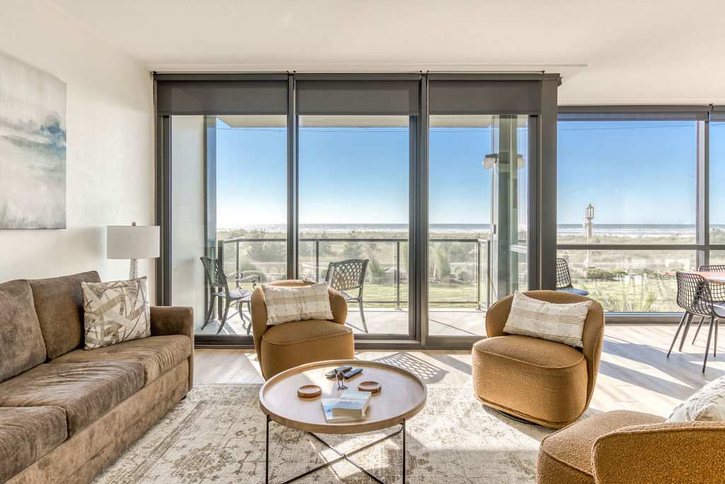 Bright living room with a brown sofa and two mustard-colored armchairs arranged around a round coffee table, facing large floor-to-ceiling windows offering expansive ocean views.