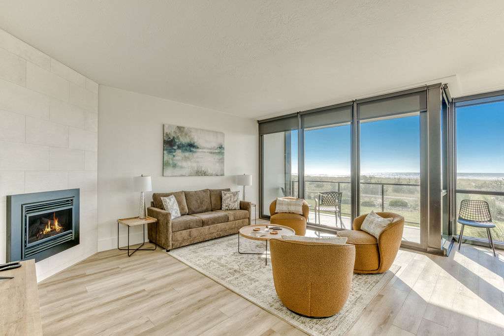 Spacious living room with a brown sofa and two mustard armchairs, centered around a fireplace and facing large windows that provide panoramic views of the ocean and coastline.