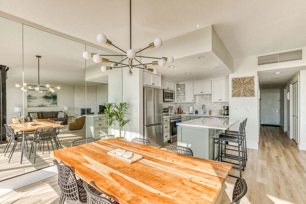 Open-concept dining area with a large live-edge wooden table and black metal chairs, illuminated by a modern sputnik chandelier. The kitchen with light cabinets and a breakfast bar with stools is visible in the background, along with a mirrored wall reflecting the living area.
