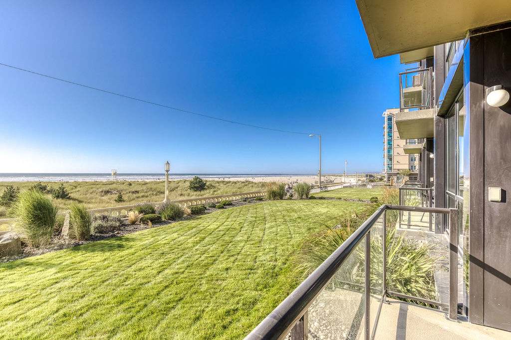 A bright, sunny view from a balcony overlooking a manicured green lawn, leading to sandy dunes and the vast ocean under a clear blue sky. A modern building with balconies is visible to the right.