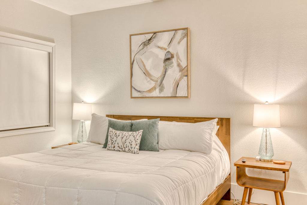 Cozy bedroom with a queen-sized bed featuring a wooden headboard, white bedding, and light blue and white decorative pillows. Two glass base lamps sit on wooden nightstands flanking the bed, with abstract framed art above the headboard.