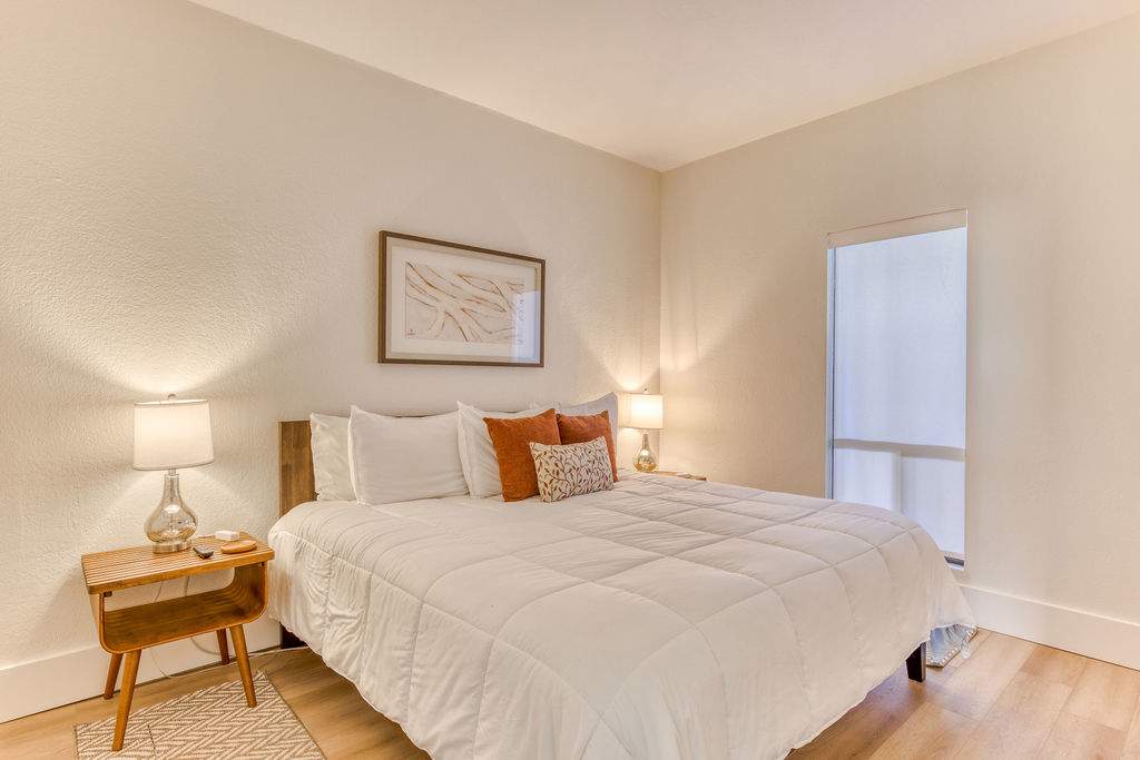 Bright bedroom with a queen-sized bed, white bedding, and orange and patterned decorative pillows. Wooden nightstands with lamps flank the bed, and a large window with a roller blind is visible on the right.