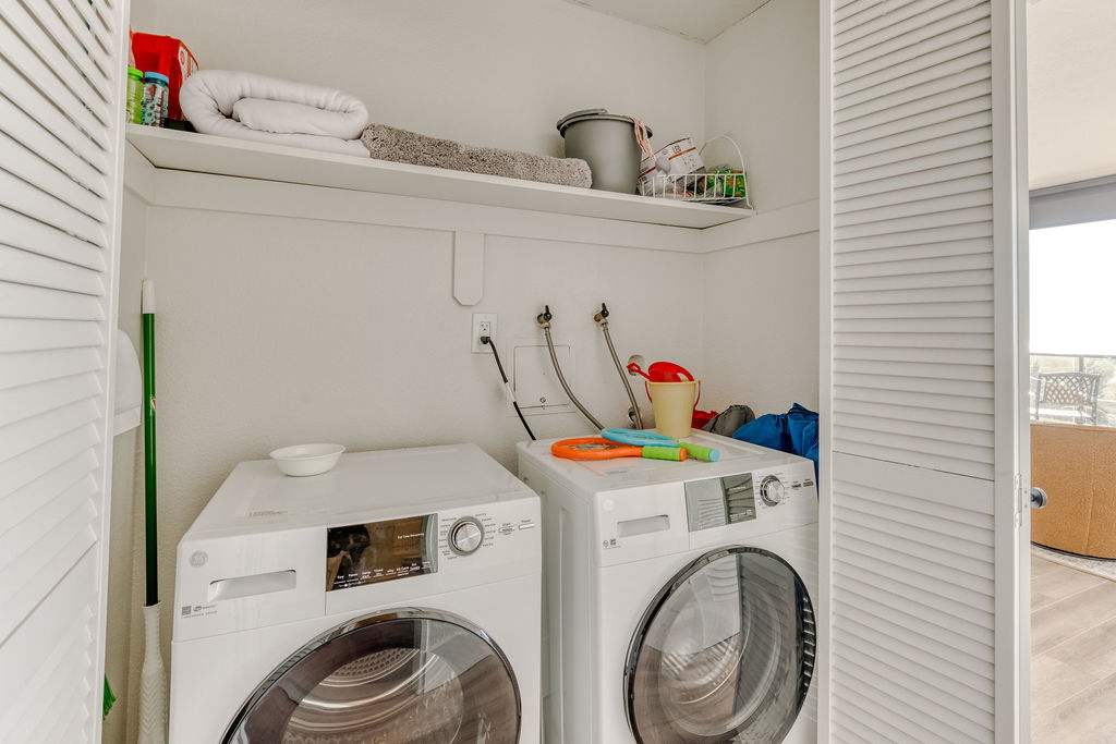 Compact laundry closet with a stackable front-loading washer and dryer, a shelf above for storage, and louvered bi-fold doors. Cleaning supplies and towels are stored on the shelf.