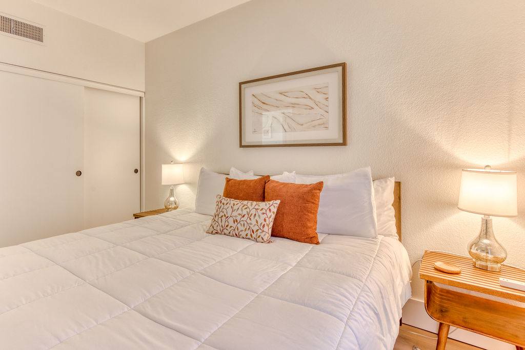 Bedroom with a queen-sized bed dressed in white bedding and accented with orange and patterned pillows. Wooden nightstands with lamps are on either side, and a sliding closet door is visible on the left.
