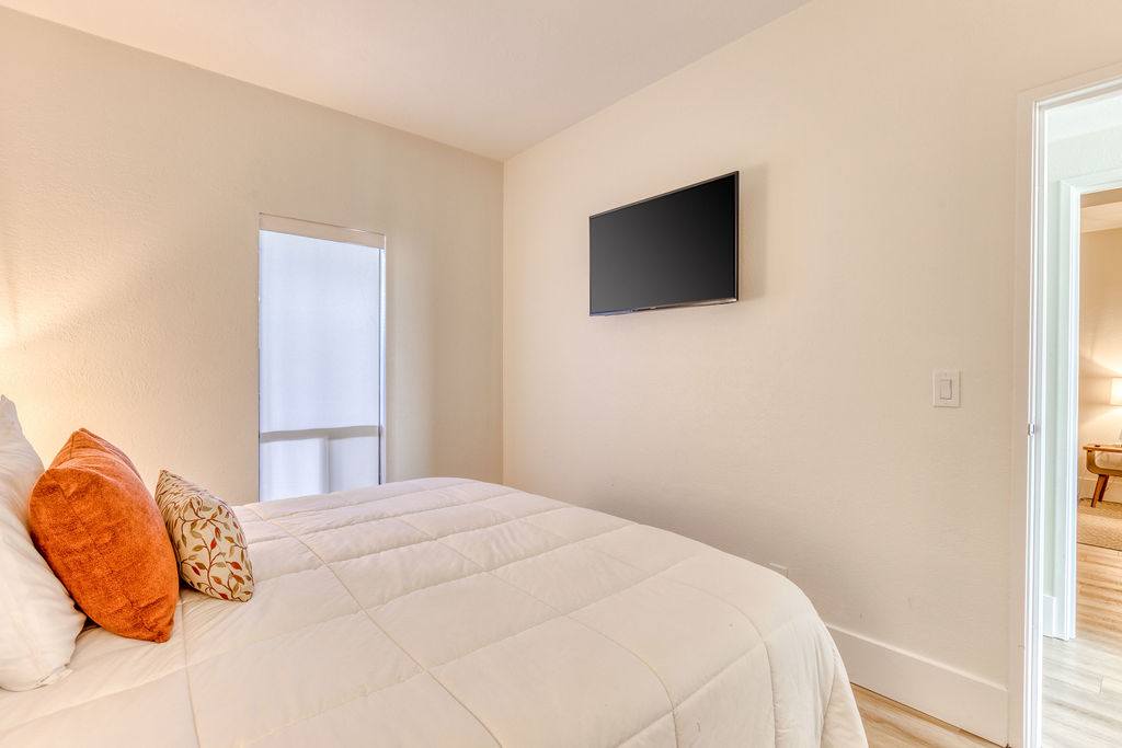 View of a bedroom wall with a wall-mounted flat-screen TV above a queen-sized bed with white bedding and orange pillows. A window with a roller blind is visible to the left of the TV.