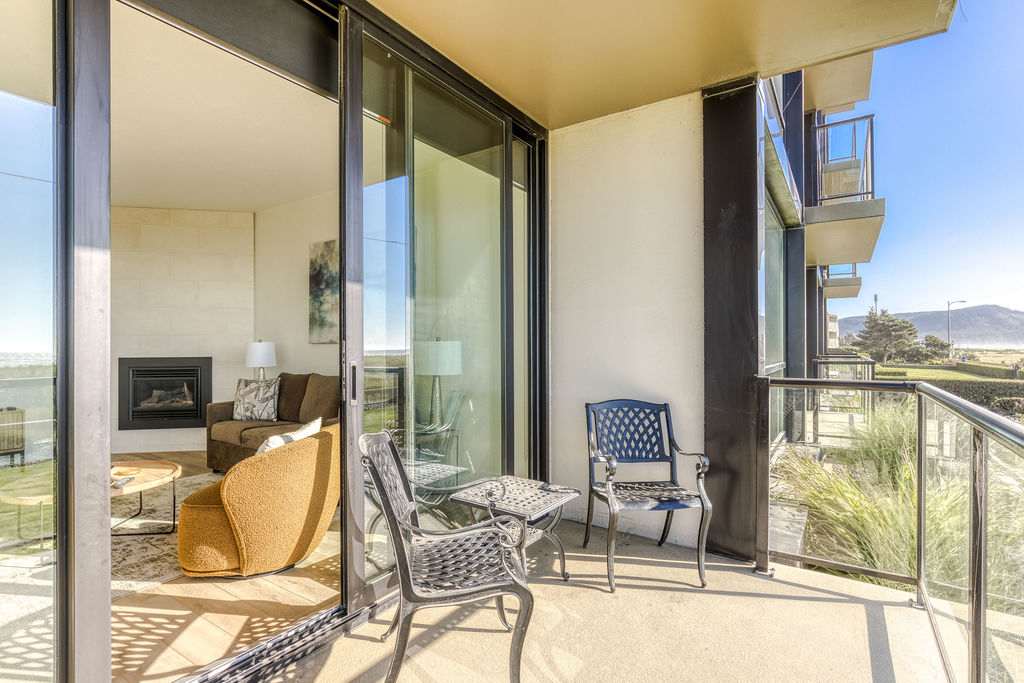 Outdoor balcony with black metal patio furniture, including two chairs and a small table, offering views of the ocean and surrounding landscape. Large sliding glass doors connect to the interior living space.