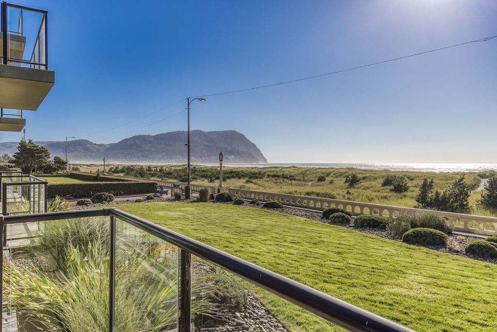 Expansive oceanfront view from a balcony, featuring a lush green lawn, sandy dunes, and the sparkling ocean with a distant mountain under a bright, sunny sky.