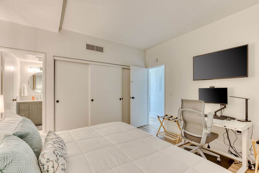 Bright bedroom featuring a queen-sized bed with white bedding, a dedicated workspace with a white desk, ergonomic chair, and monitor, and a wall-mounted flat-screen TV. A sliding closet door is visible, and a bathroom entrance is to the left.