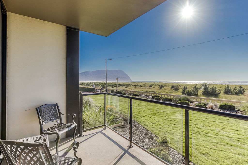 A cozy balcony furnished with two black metal chairs and a small table, offering a stunning oceanfront view of green lawns, sand dunes, and the sparkling sea under a bright, sunny sky.