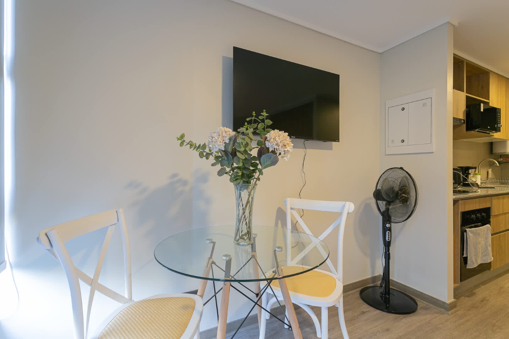 Cozy dining nook with a glass table, perfect for intimate meals, complemented by a wall-mounted TV and a stylish kitchen corner.