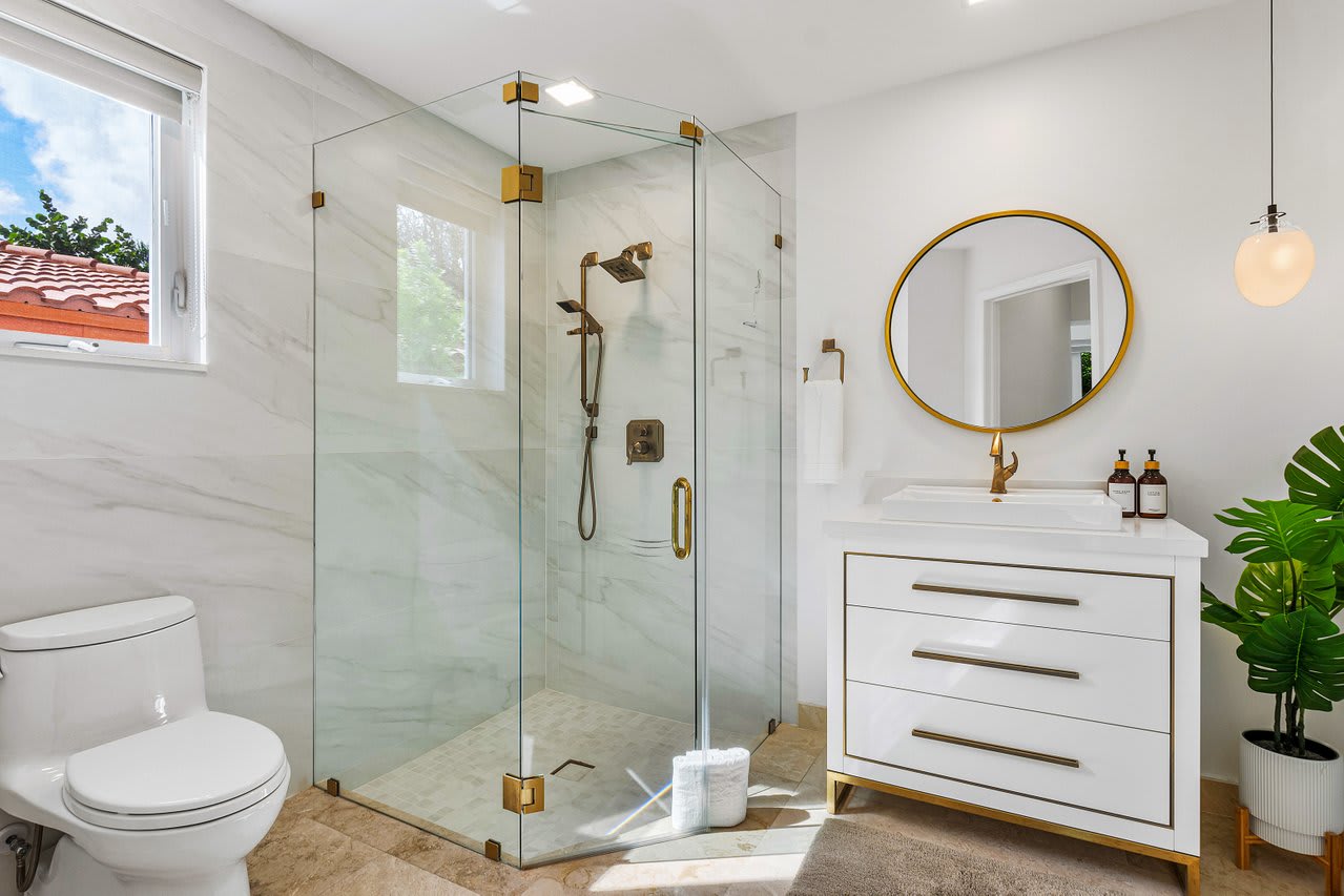 Indulge in elegance with a glass-enclosed rain shower, brass fixtures, and a chic marble vanity, set against crisp white walls.