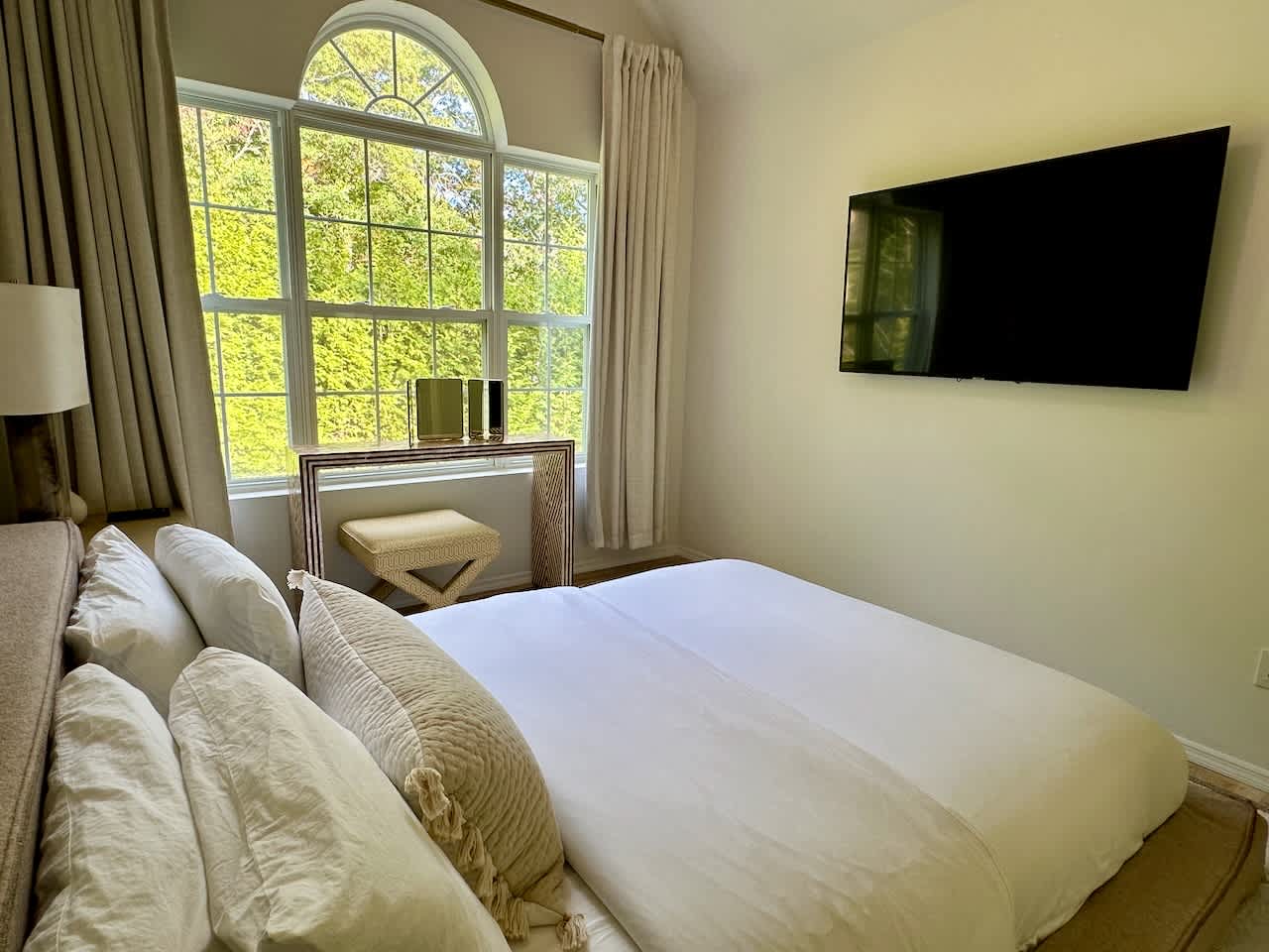 Bedroom with a queen bed, wall-mounted TV, and an arched window overlooking greenery.