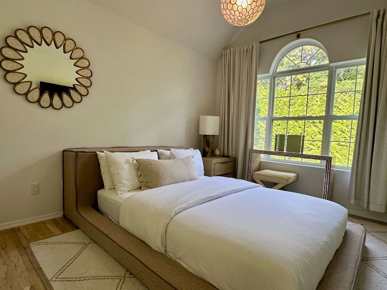 Bedroom with a wood-framed bed, round sunburst mirror, and a garden-view arched window.