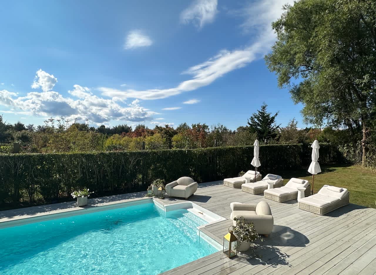 Pool deck with lounge chairs and umbrellas set against a hedge-lined lawn under a blue sky.