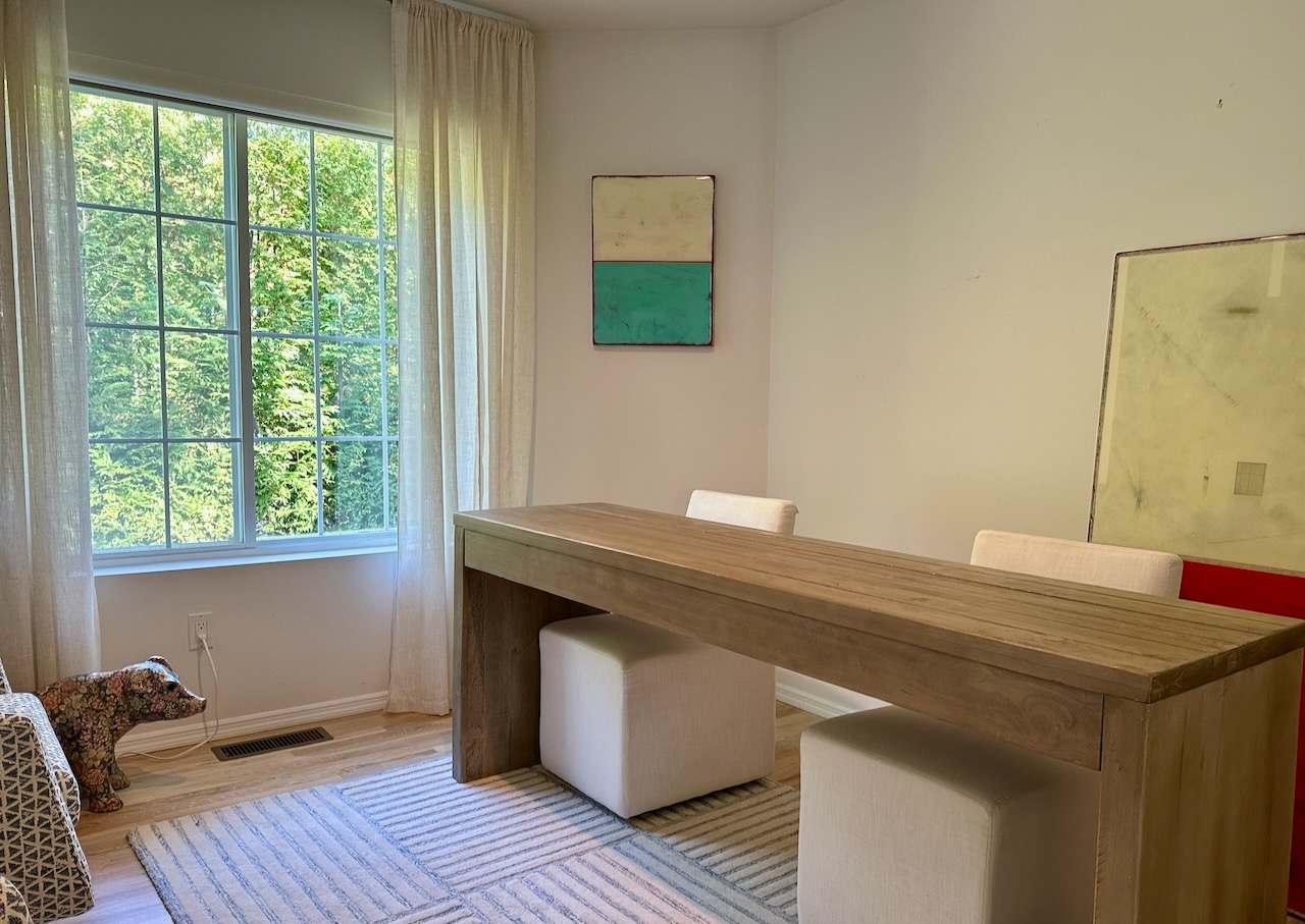 Den with a wood console table, stools, and abstract artwork by a garden-view window.