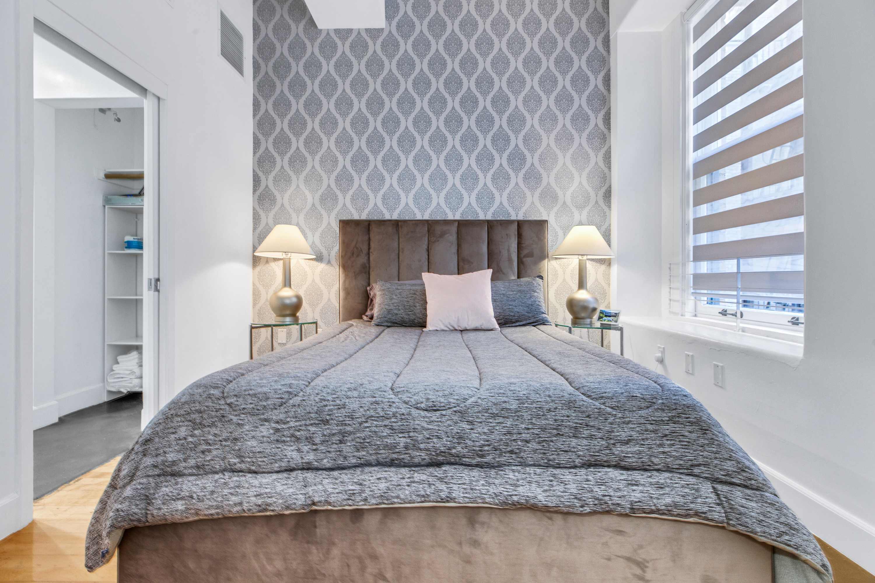 A neatly made bed with a gray comforter and pillows in a room with a window, closet, and patterned wallpaper.