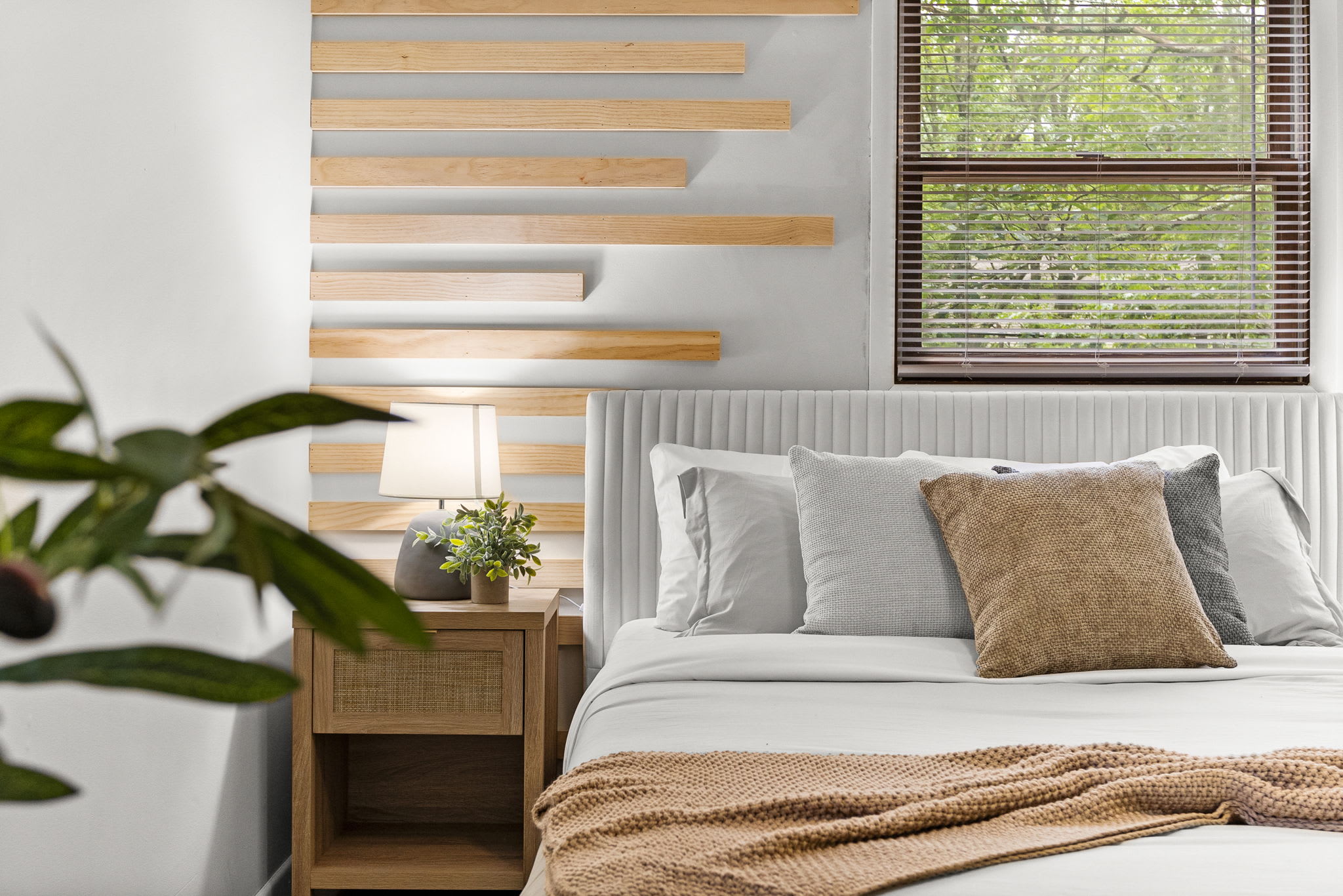 Another perspective highlighting the bedside nightstand, indoor plants, and large windows allowing natural light.