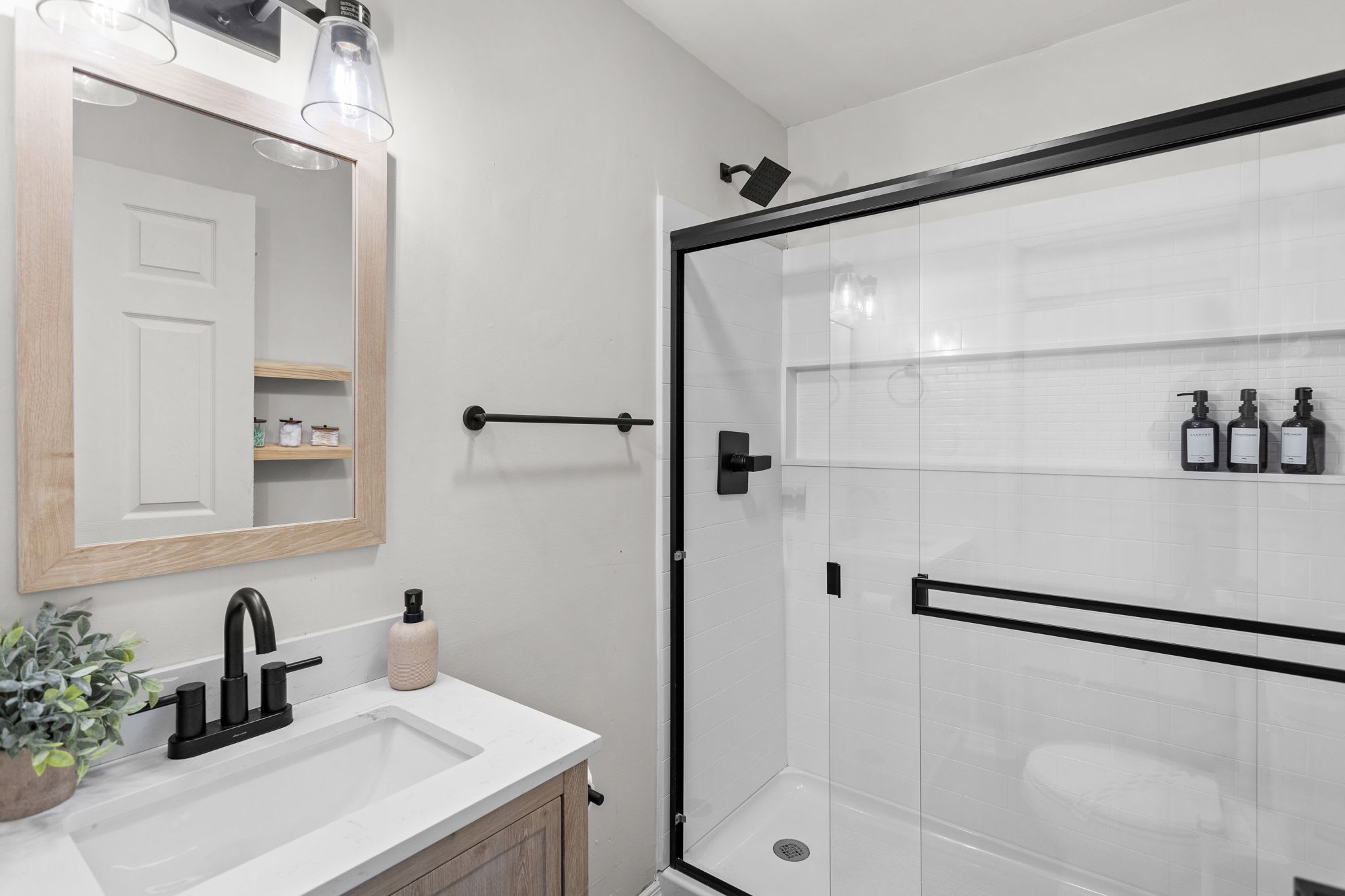 Sleek Bathroom Design – The black-framed shower glass, matte black fixtures, and mounted shampoo dispensers add a contemporary touch.