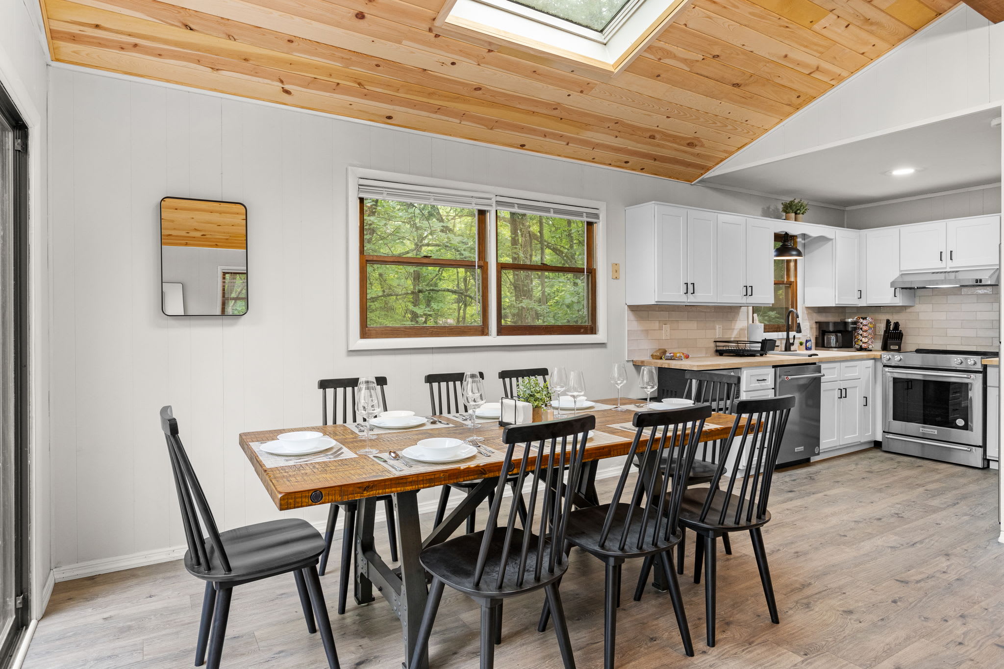 A long wooden dining table with black modern chairs sits beneath a skylight, giving the space an airy, open feel.