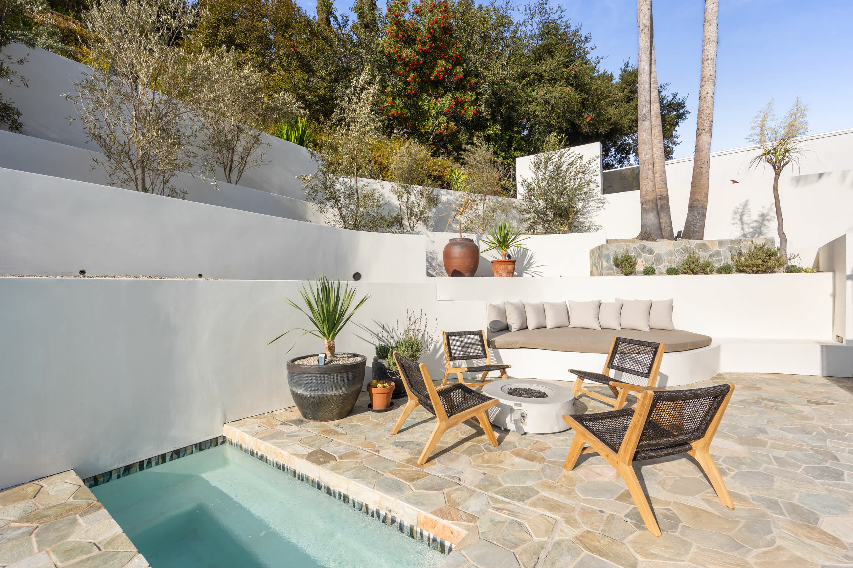 Sunlit patio with a sunken hot tub, natural wood chairs, and layered greenery.