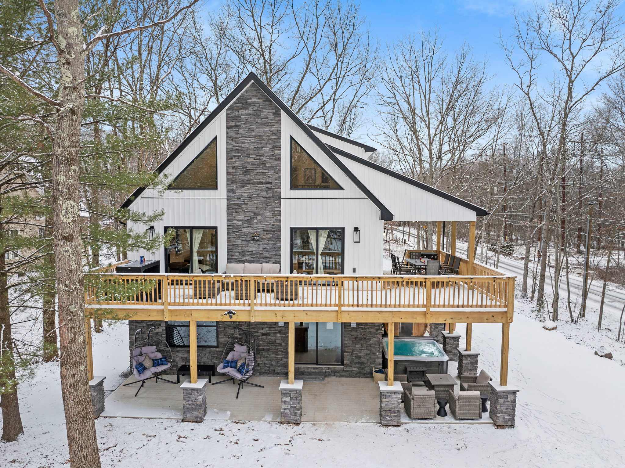 A modern A-frame cabin surrounded by snow-covered trees, complete with an expansive deck and a private hot tub. The perfect escape in nature!