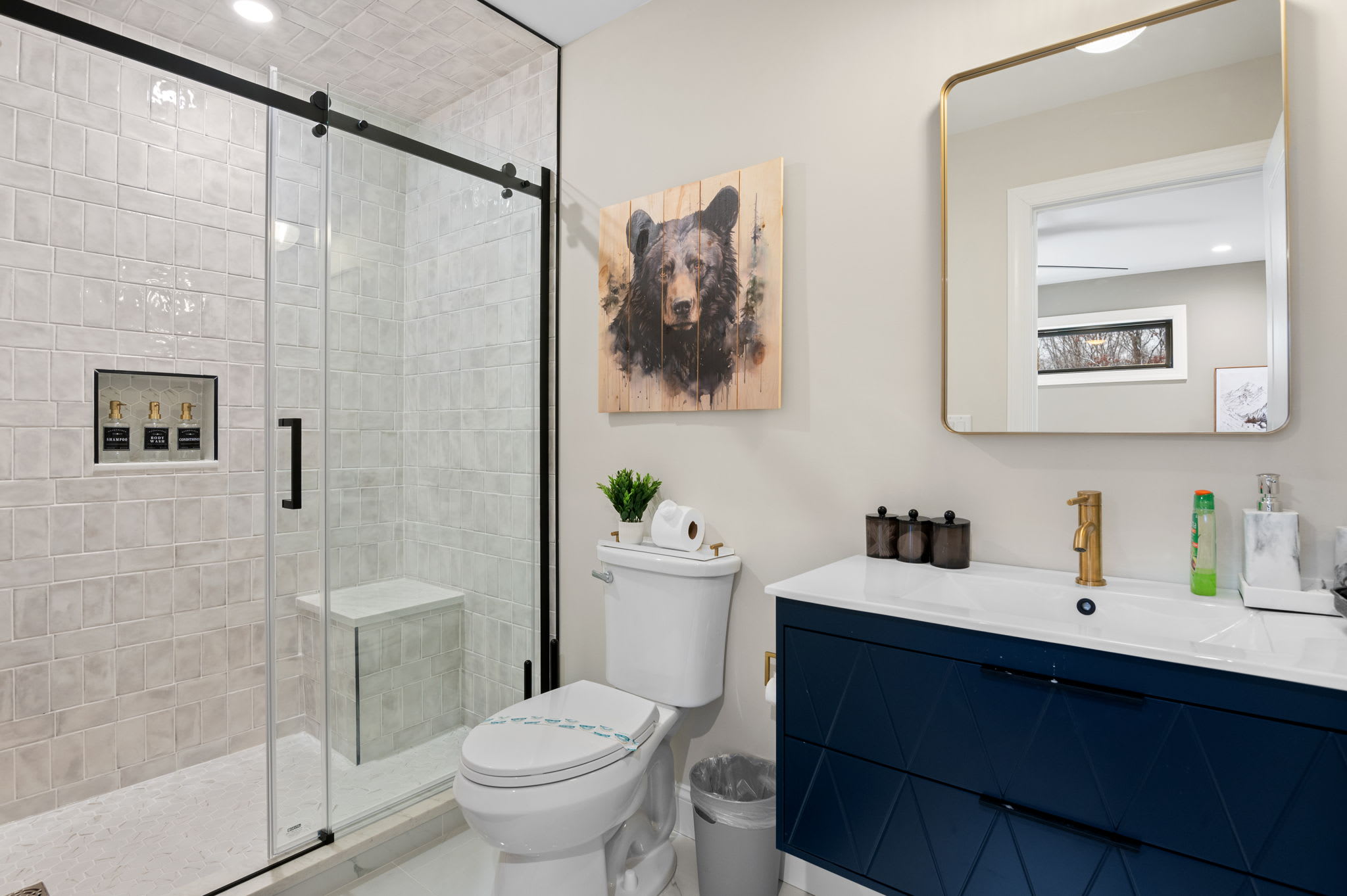 A sleek glass shower, bold navy vanity, and gold fixtures bring a modern edge to the bathroom’s rustic bear-themed art.