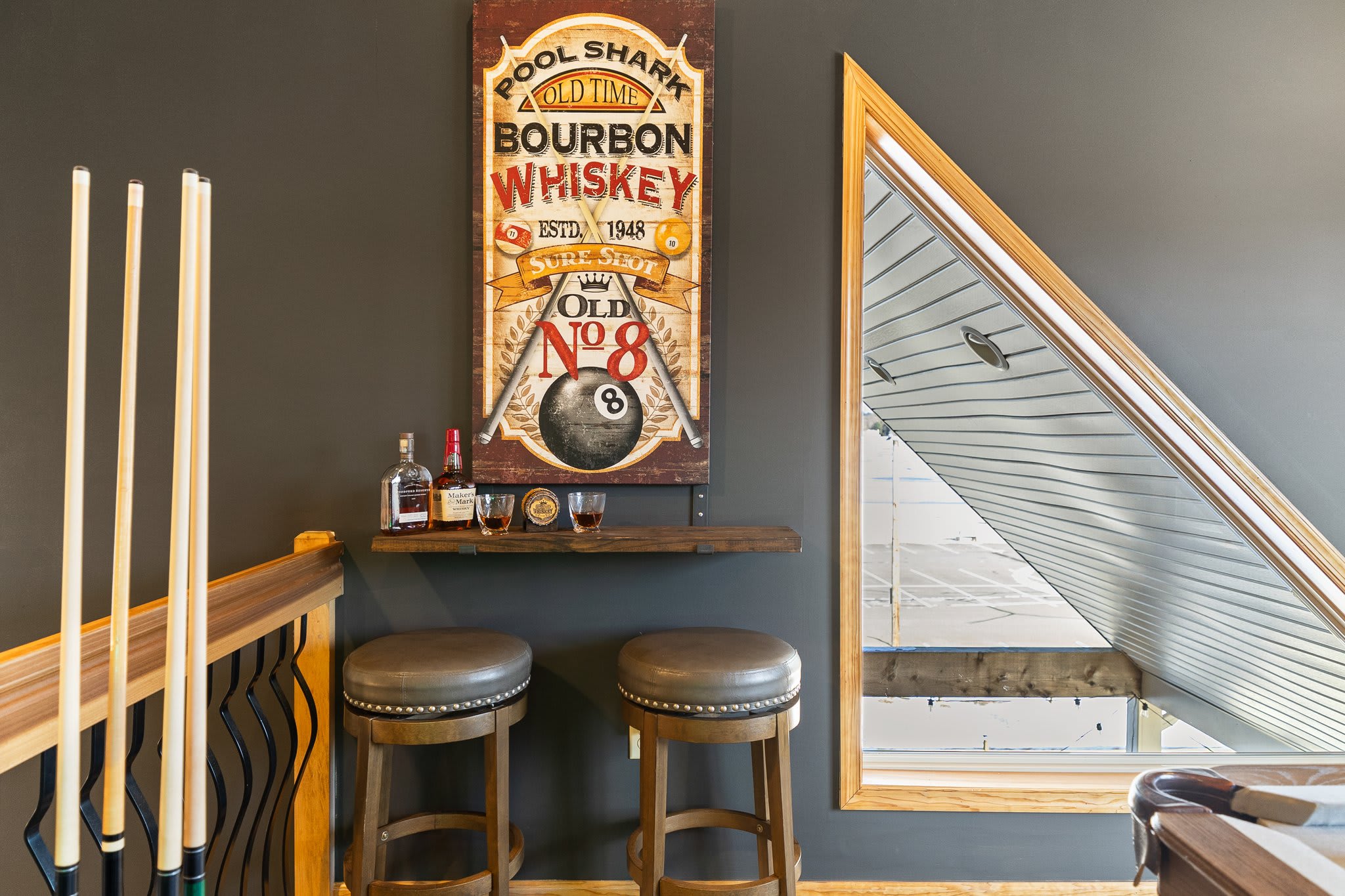 A cozy little bar corner under the stairs with stools and a vintage whiskey sign, pool cue storage nearby.