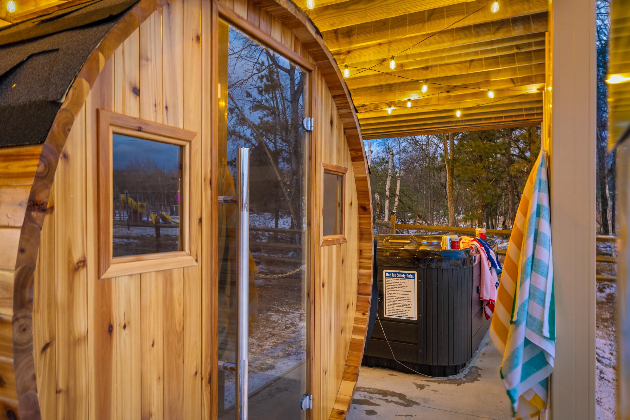 A gorgeous cedar barrel sauna with string lights, towels ready to go, and a hot tub peeking out behind, your wellness escape starts here.