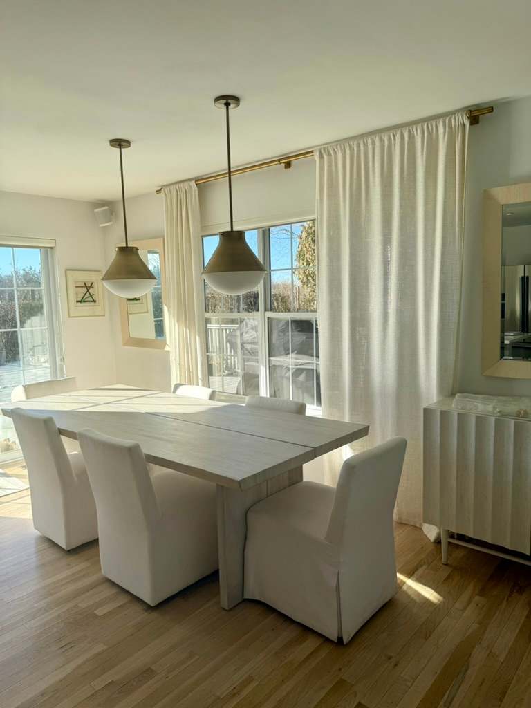 Dining area with a white table and chairs beneath brass pendant lights, near the kitchen.