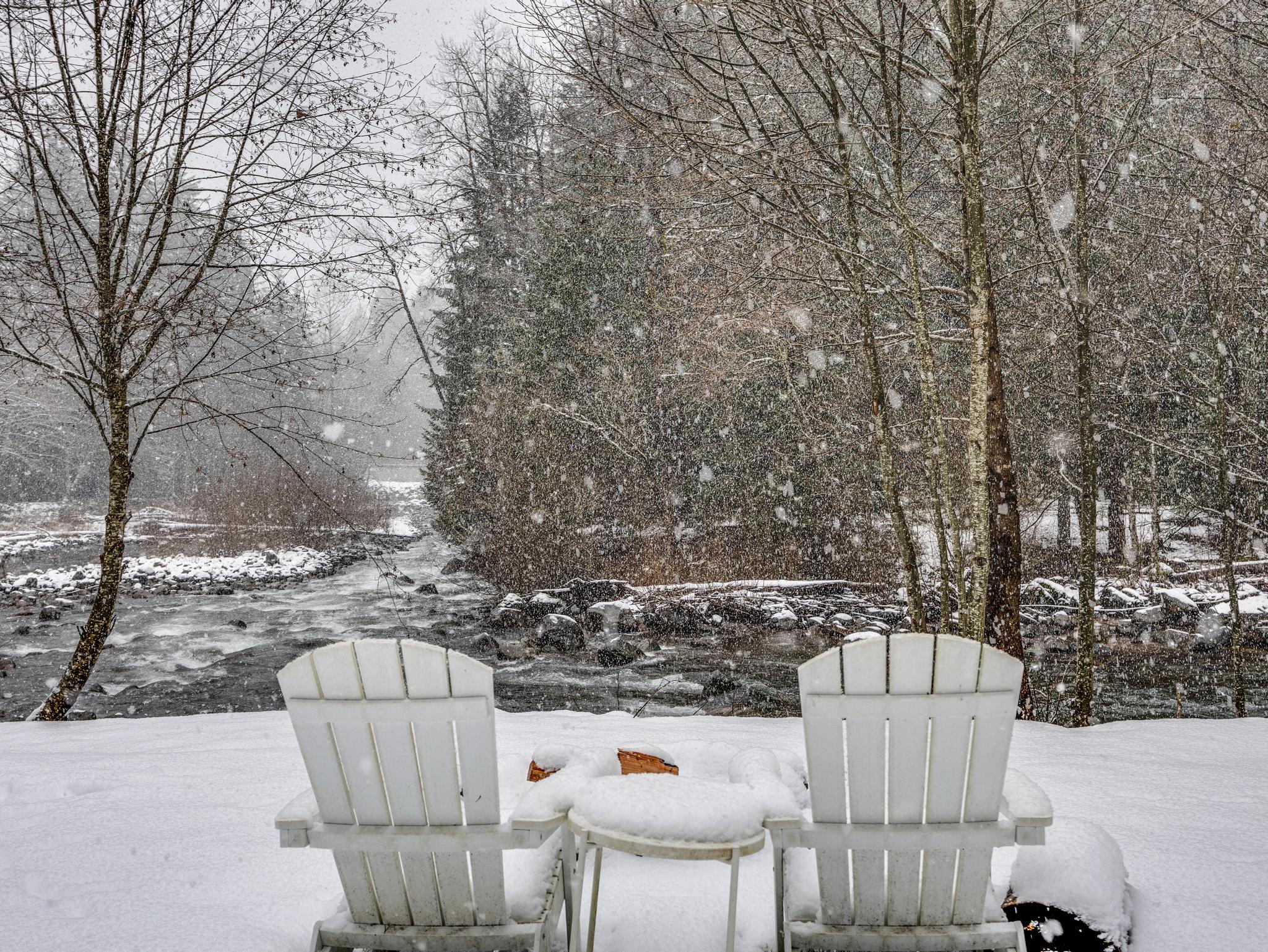 Two white Adirondack chairs and a small table, all covered in snow, positioned in a snowy yard facing a flowing river surrounded by snow-dusted trees during a winter snowfall.