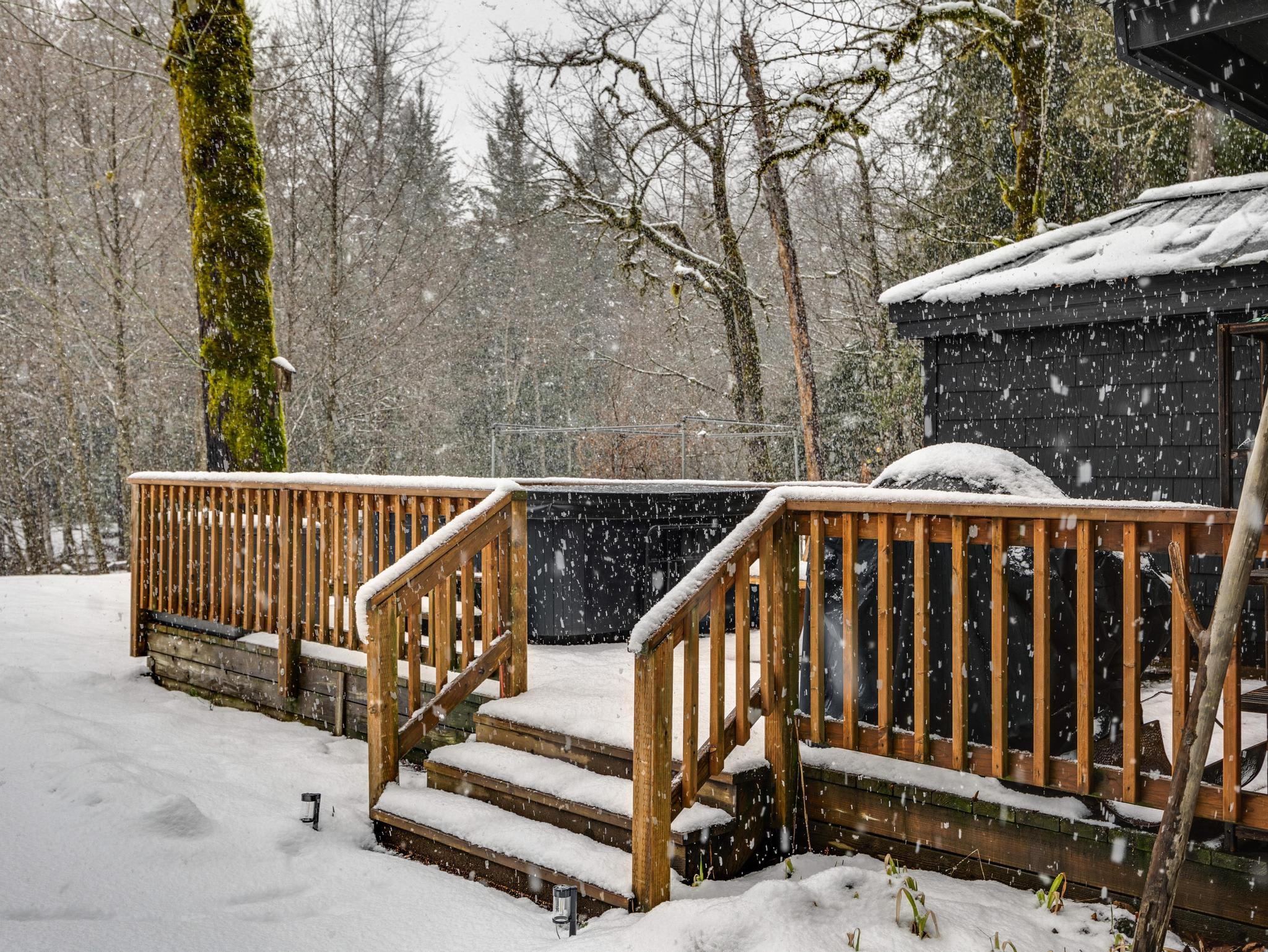 A wooden deck covered in fresh snow, featuring a dark hot tub and a wooden railing, surrounded by snow-dusted trees during a light snowfall.