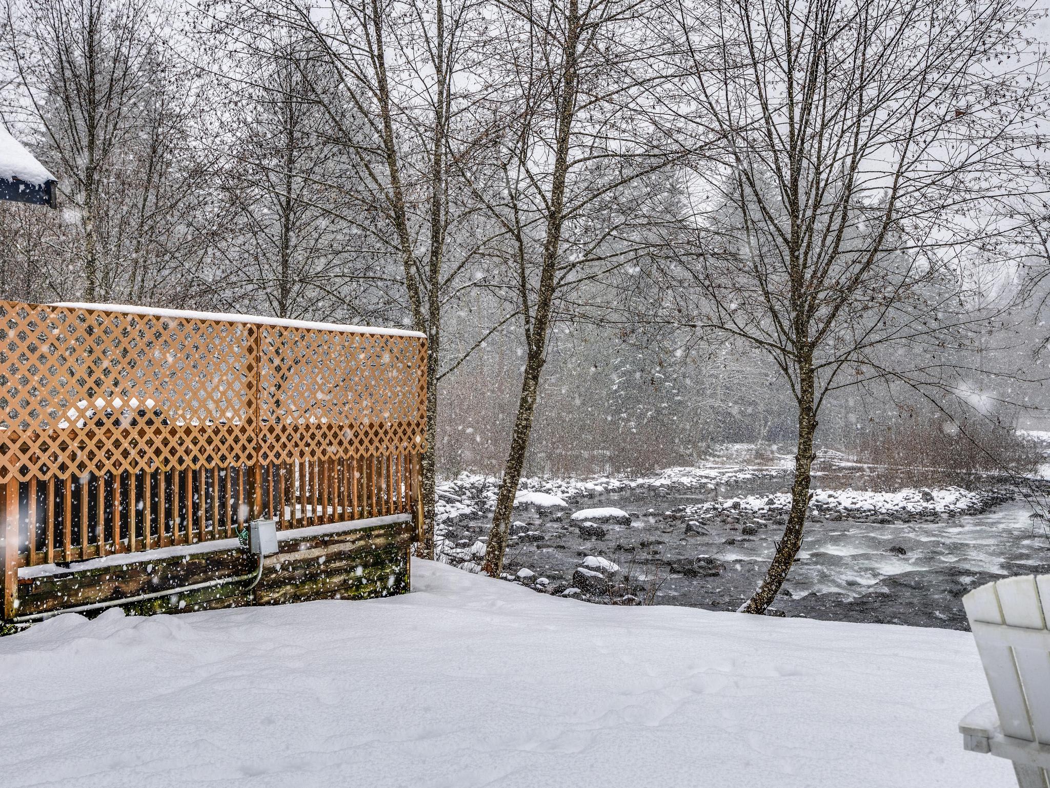 A snow-covered wooden deck with a lattice railing, overlooking a rocky river and bare trees, with snow falling gently in the background.