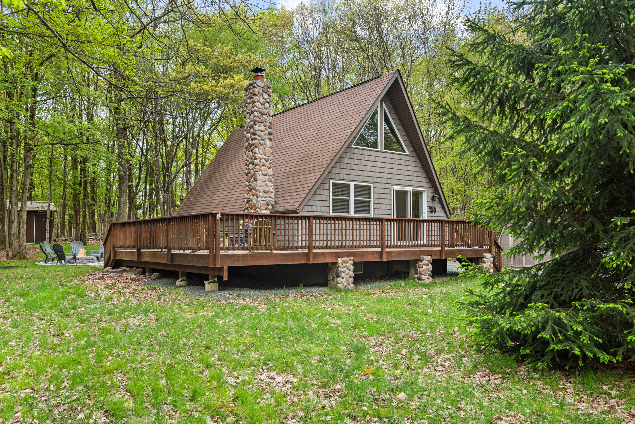 A picturesque front view of the A-frame cabin, showcasing its beautiful architecture, spacious wooden deck, and natural stone accents. Surrounded by vibrant summer foliage, it's the perfect setting for a cozy getaway.