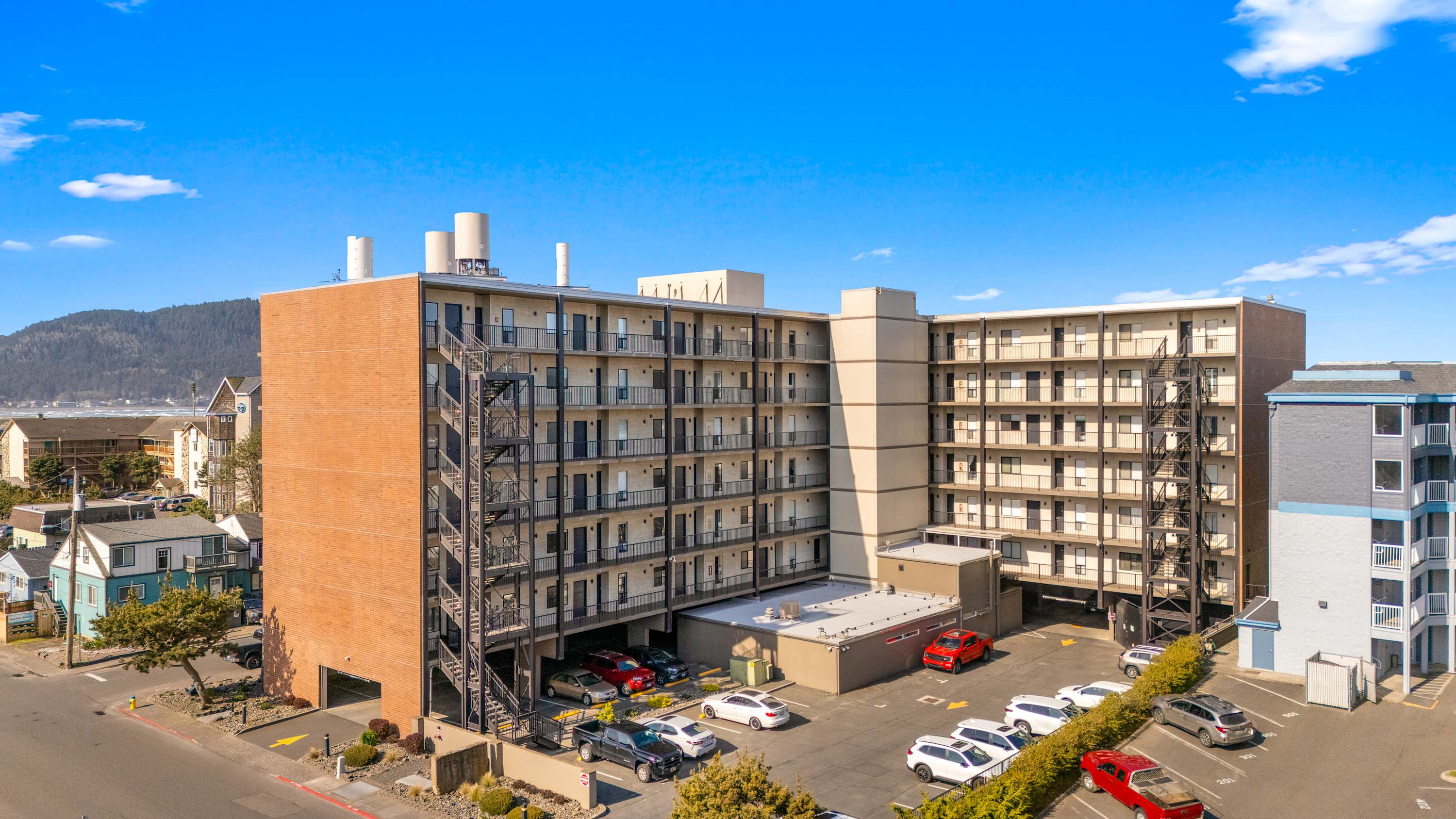 An aerial view showcasing a large, multi-story condominium building with brick and light-colored facades, numerous balconies, and external staircases, surrounded by parking areas with the ocean and mountains in the distance.