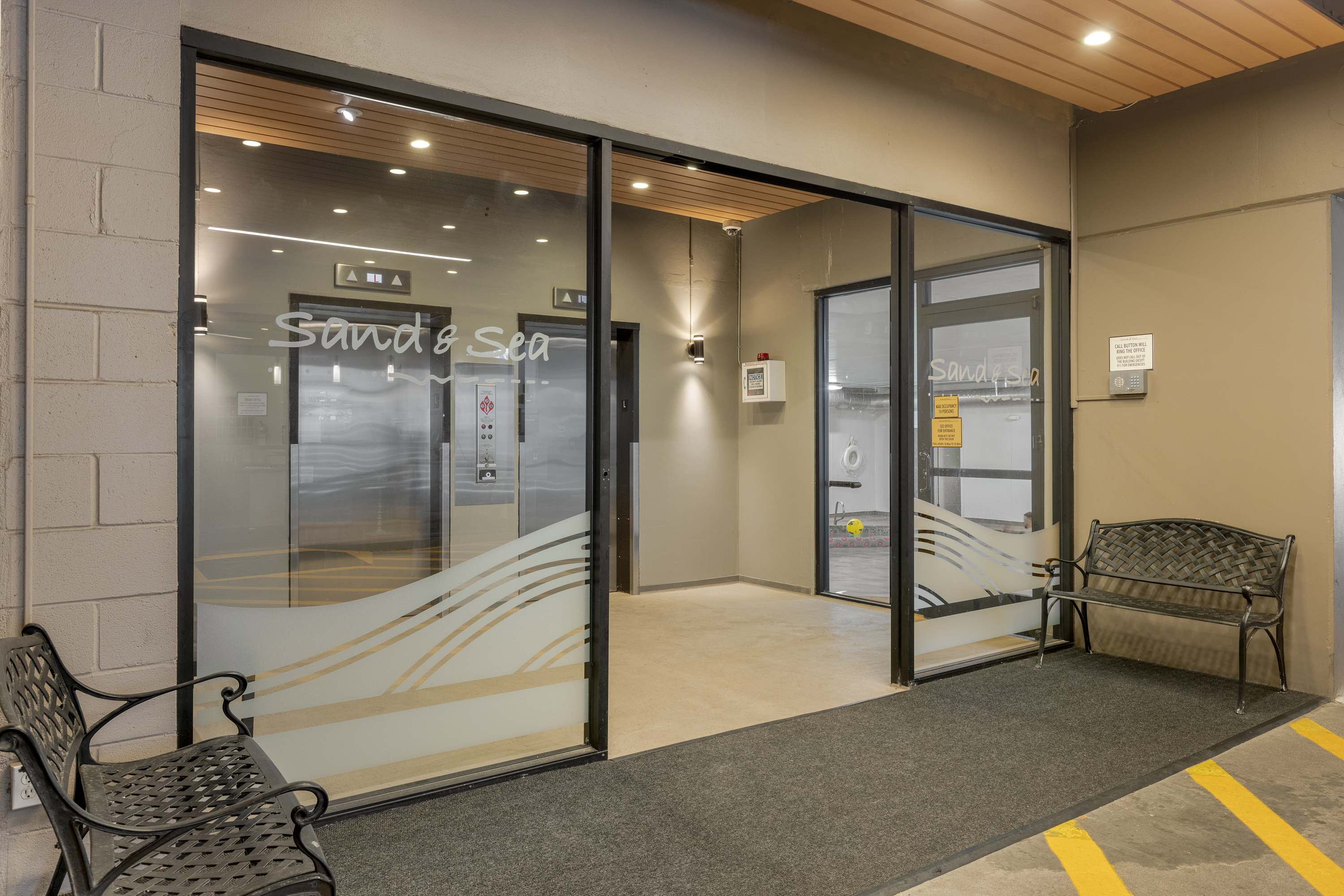 Modern building entrance with large glass doors featuring frosted wave patterns and "Sand & Sea" branding, leading to elevators. A black metal bench sits outside on the left.