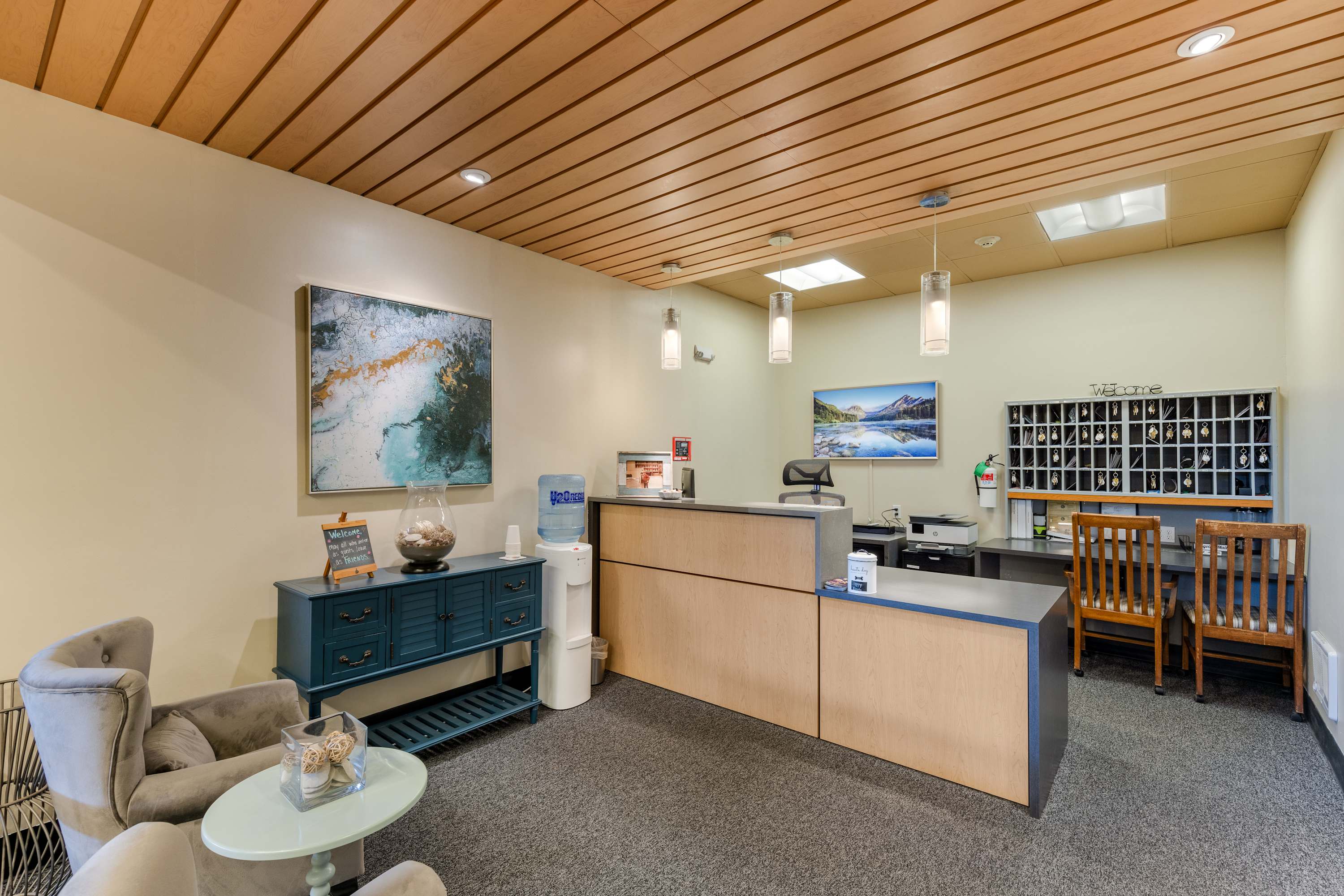 Bright and welcoming reception area with a light wood slatted ceiling, a modern reception desk, and a water cooler. Abstract art hangs on the wall, and two pendant lights illuminate the space.